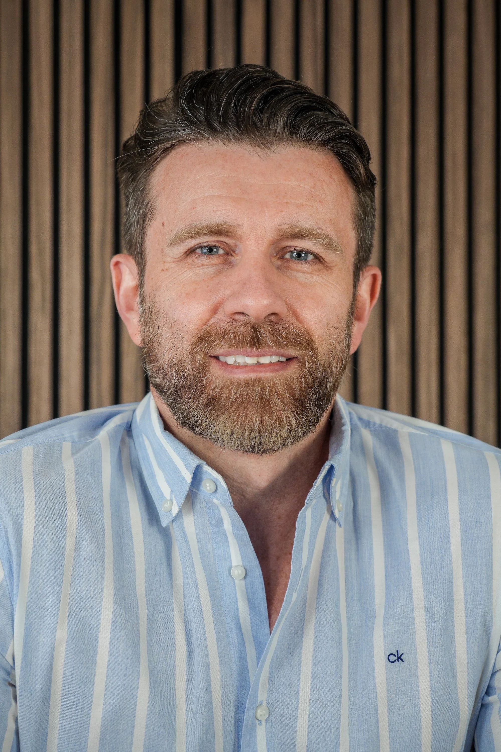 A man with a beard and blue eyes, wearing a light blue striped button-up shirt, smiling in front of a wooden panel background.