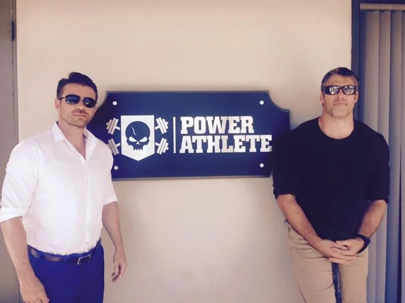 Two men wearing sunglasses stand on either side of a sign that says "Power Athlete" with a skull and barbell graphic.