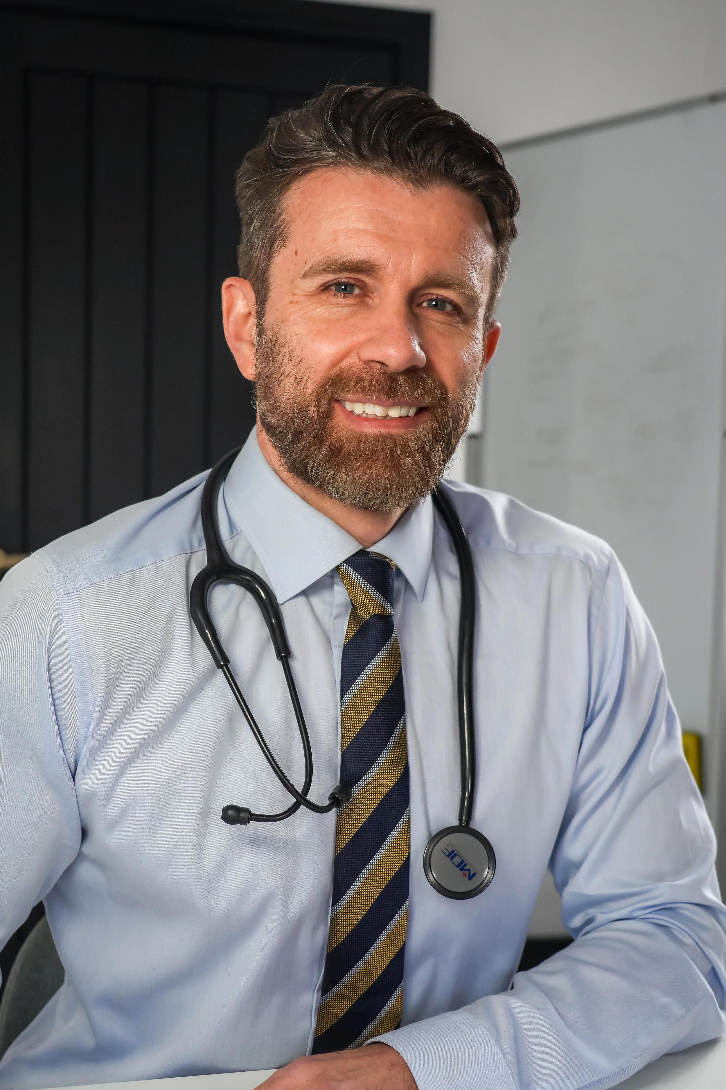 A male doctor with a beard and mustache, wearing a light blue dress shirt, patterned tie, and stethoscope around his neck, sitting indoors and smiling at the camera.