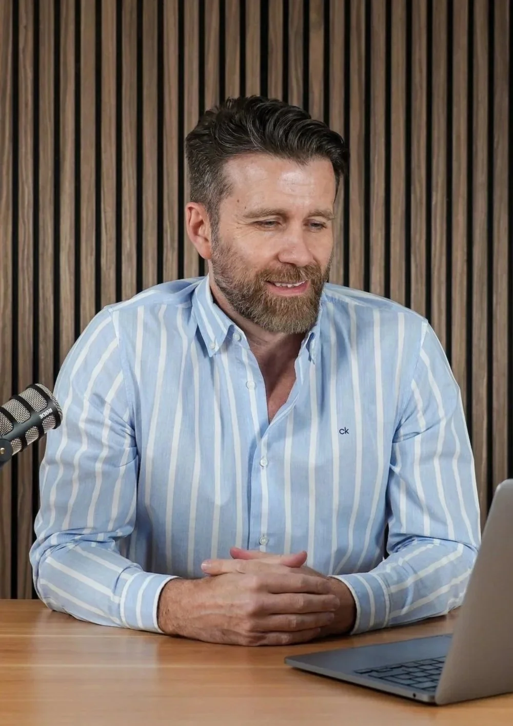 A man with a beard and styled hair, wearing a light blue and white striped shirt, sitting at a wooden table in front of a laptop, with a microphone nearby, in front of a wooden slatted wall.