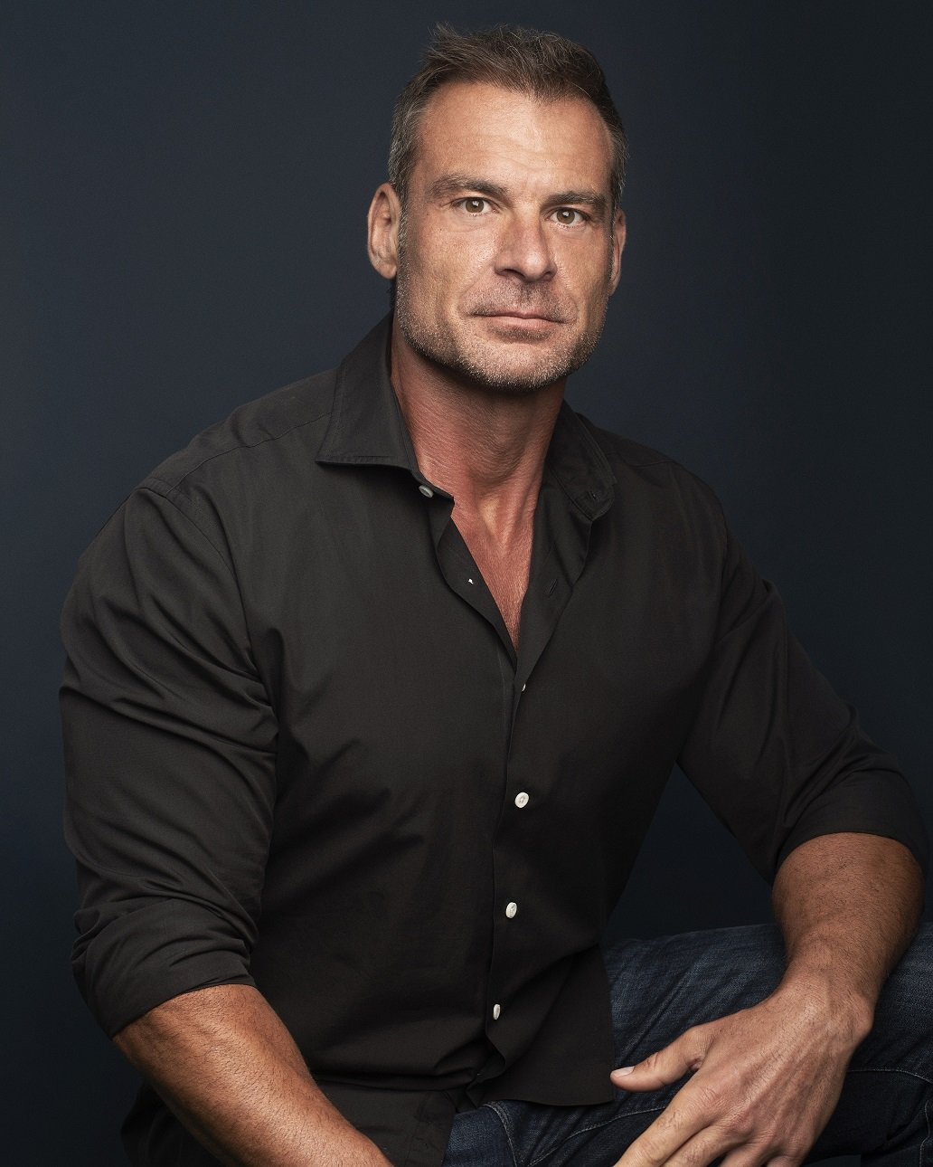 A portrait of a middle-aged man with short brown hair and stubble, wearing a black button-up shirt with the sleeves rolled up, sitting against a dark background.