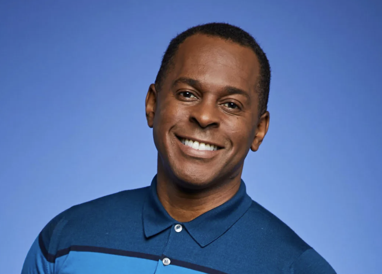 A smiling man with short hair wearing a blue collared shirt against a blue background.