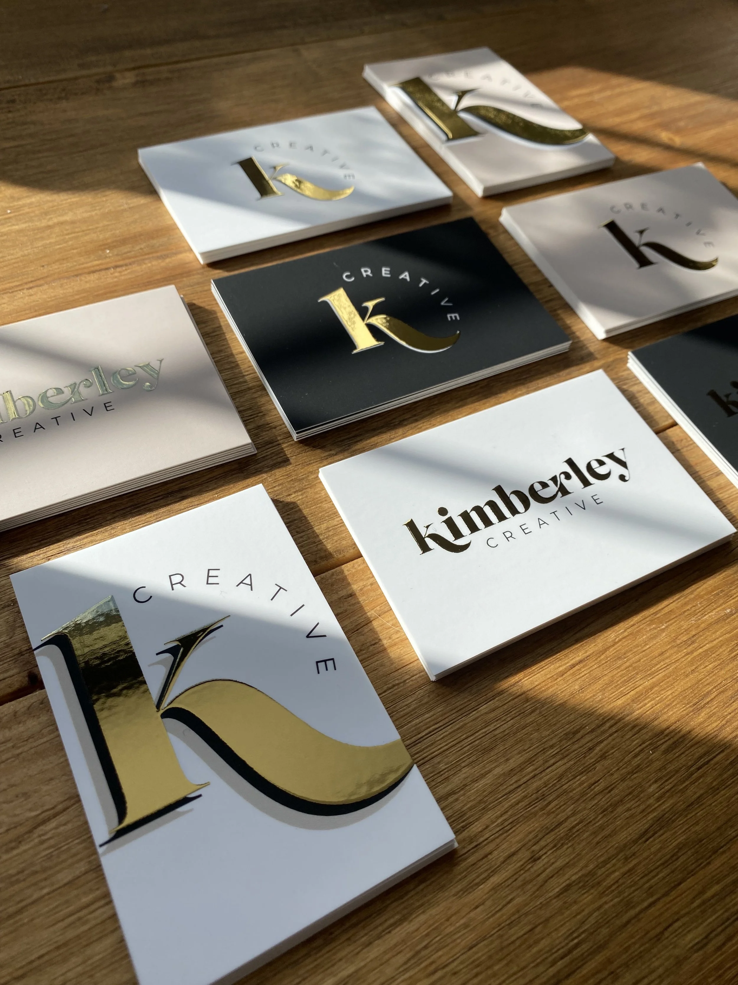 Collection of business cards with "Kimberley Creative" branding, featuring the letter 'K' in gold foil and various color backgrounds on a wooden surface.
