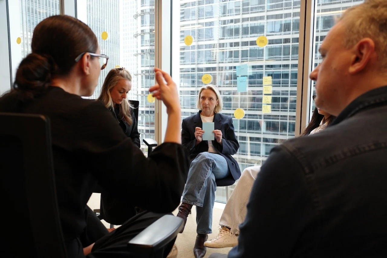 A group of five people in a corporate meeting room with large glass windows, with a cityscape of tall buildings in the background. One woman is sitting in the center holding a sticky note, while others listen attentively.