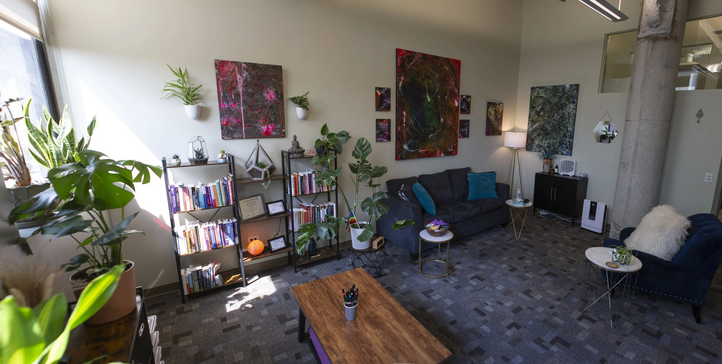 Cozy living room with black couch, blue armchair, and artwork on the walls; includes plants, bookshelves, side tables, and a lamp.