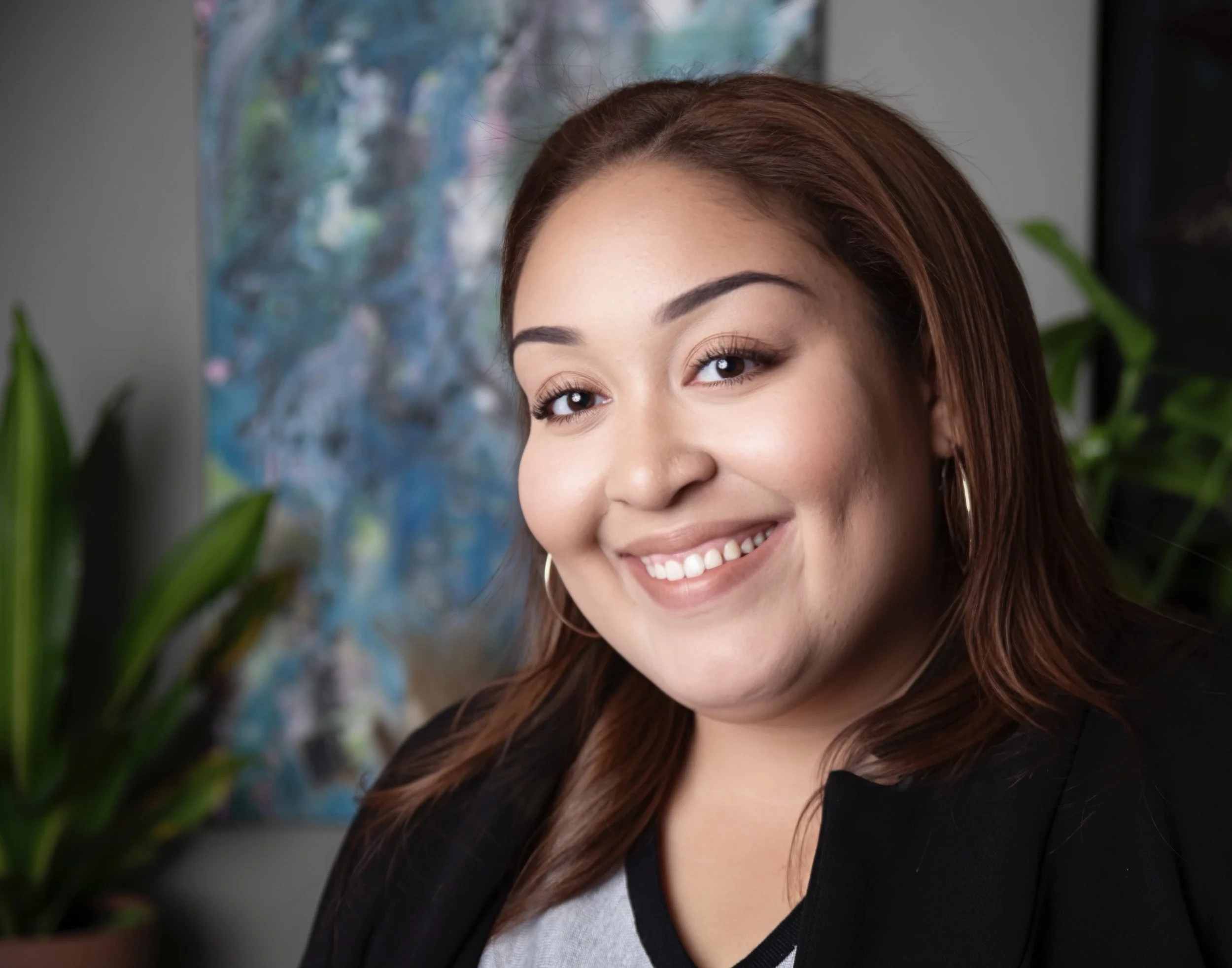 Smiling young woman with brown hair, wearing hoop earrings and a black blazer, standing indoors near a colorful abstract painting and green plants.