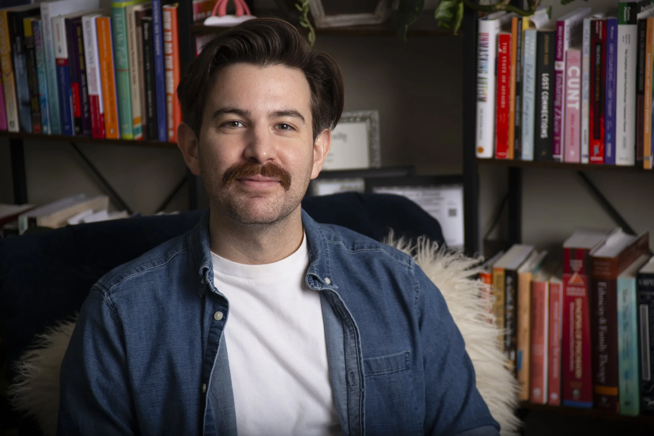 A man with dark brown hair, light skin, and a beard, smiling at the camera, wearing a black shirt, against a plain beige background.