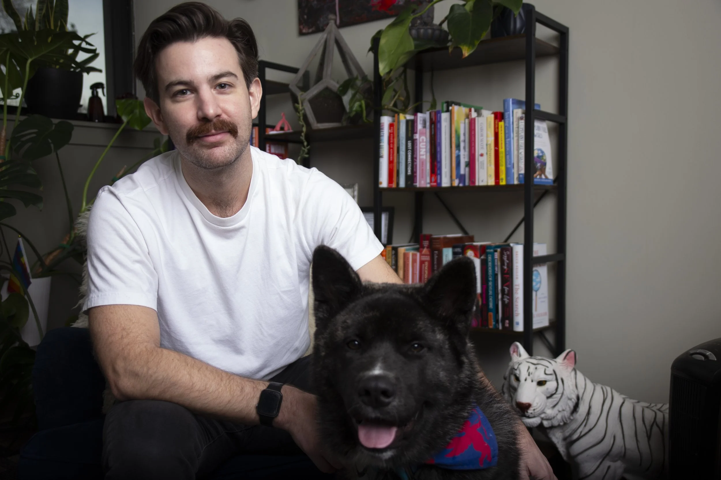 A man with brown hair, mustache, wearing a white t-shirt and black watch, sitting next to a black and white dog wearing a blue bandana, with a white tiger plush toy in the background, in a room with bookshelves and plants.