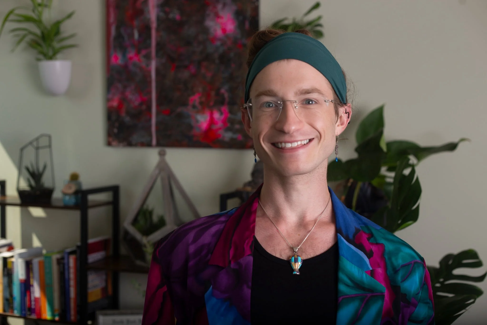 A smiling person with light skin, wearing glasses, a black headband, and a black and white striped shirt with yellow accents, sitting on a blue chair with a white fluffy pillow.