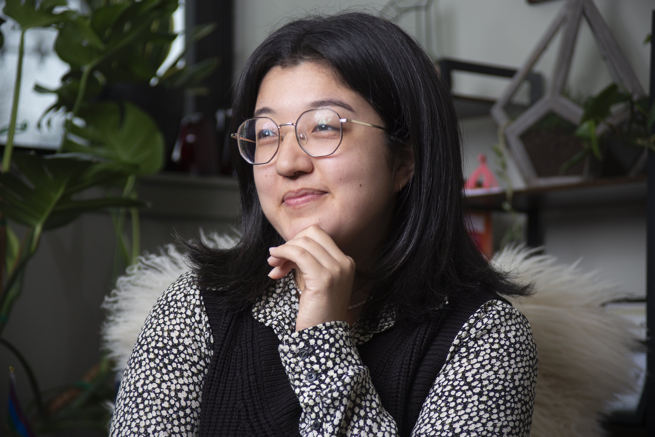 A woman with black hair and glasses sitting thoughtfully in an indoor space filled with green plants and modern decor.
