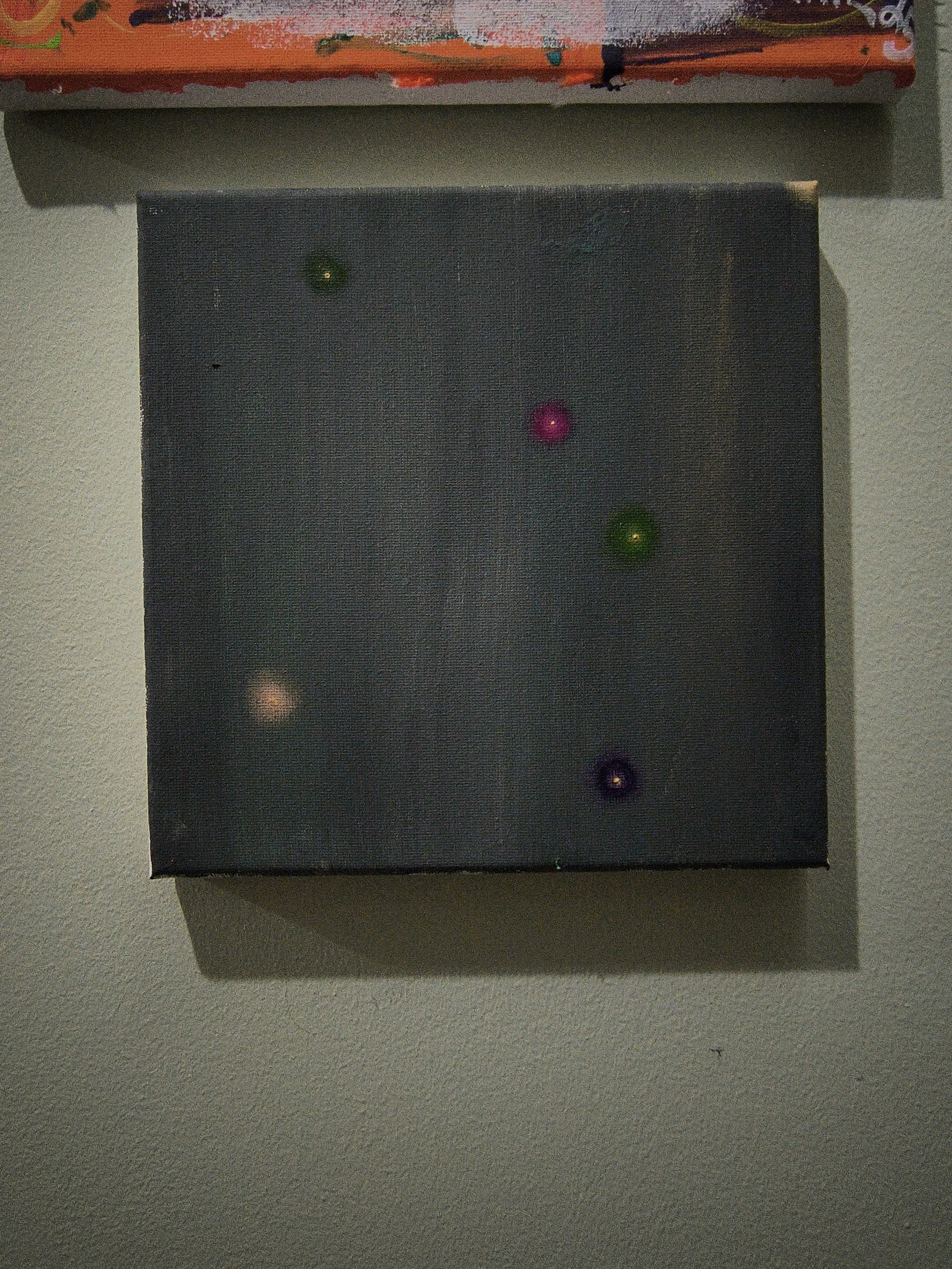A black square canvas with small, colorful dots attached, hung on a wall.