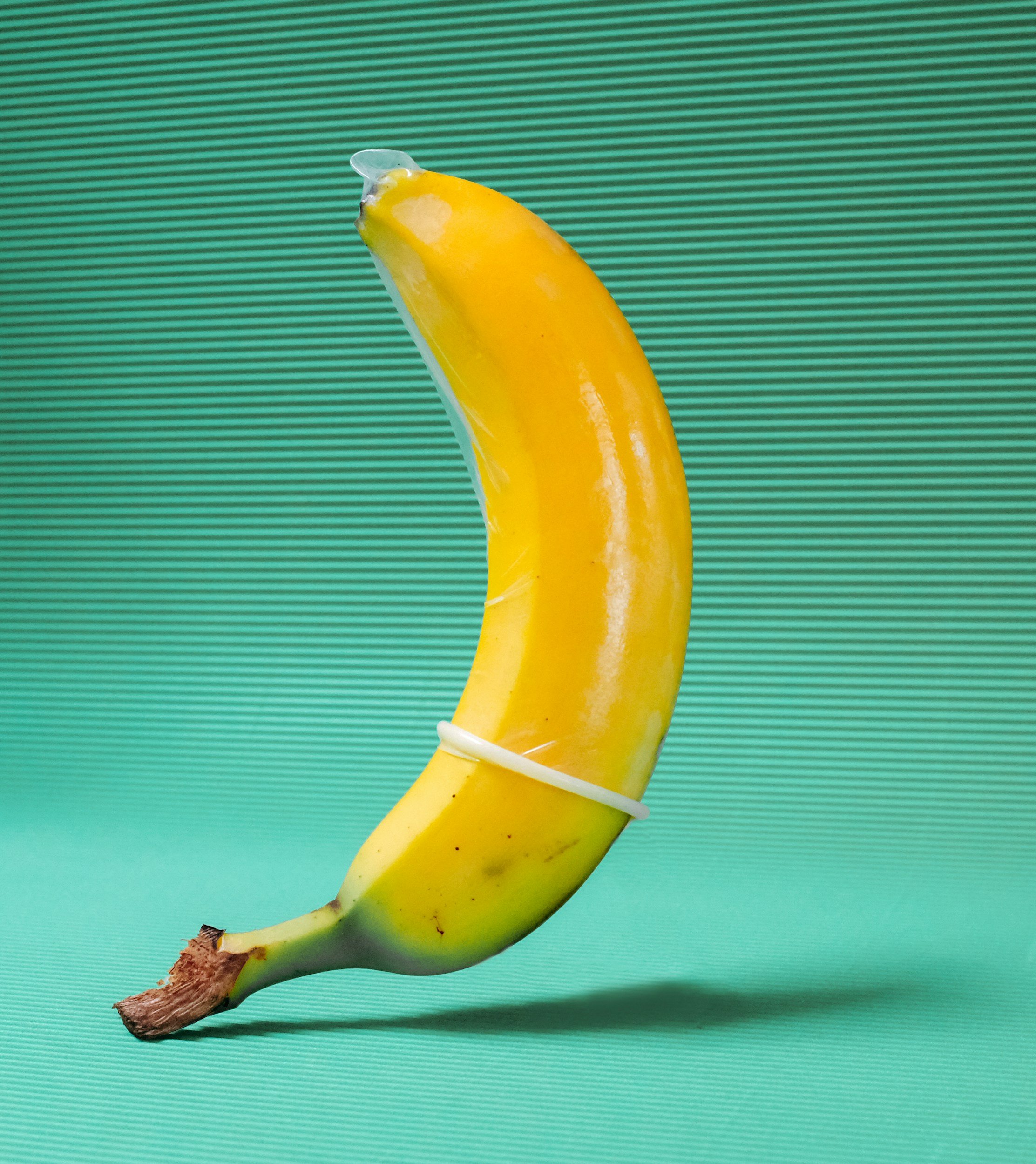 A banana with a plastic covering around it, shaped to look like a crescent moon, placed on a teal surface with a teal striped background.