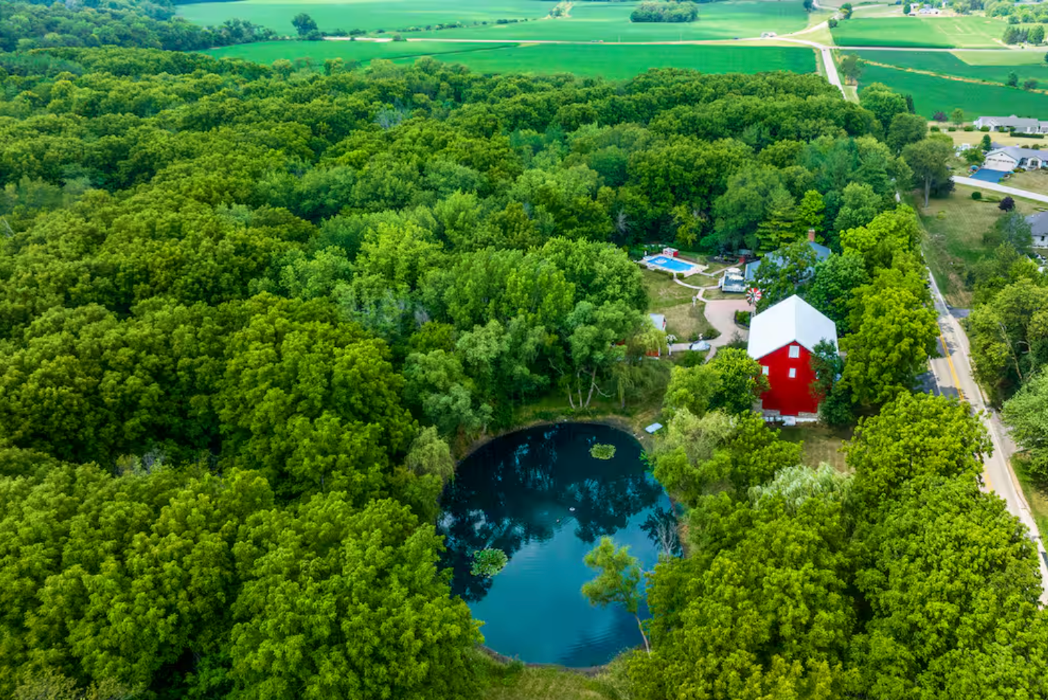 Aerial view of a lush green forest with a small pond, a red barn, and a swimming pool nearby, surrounded by fields and roads.