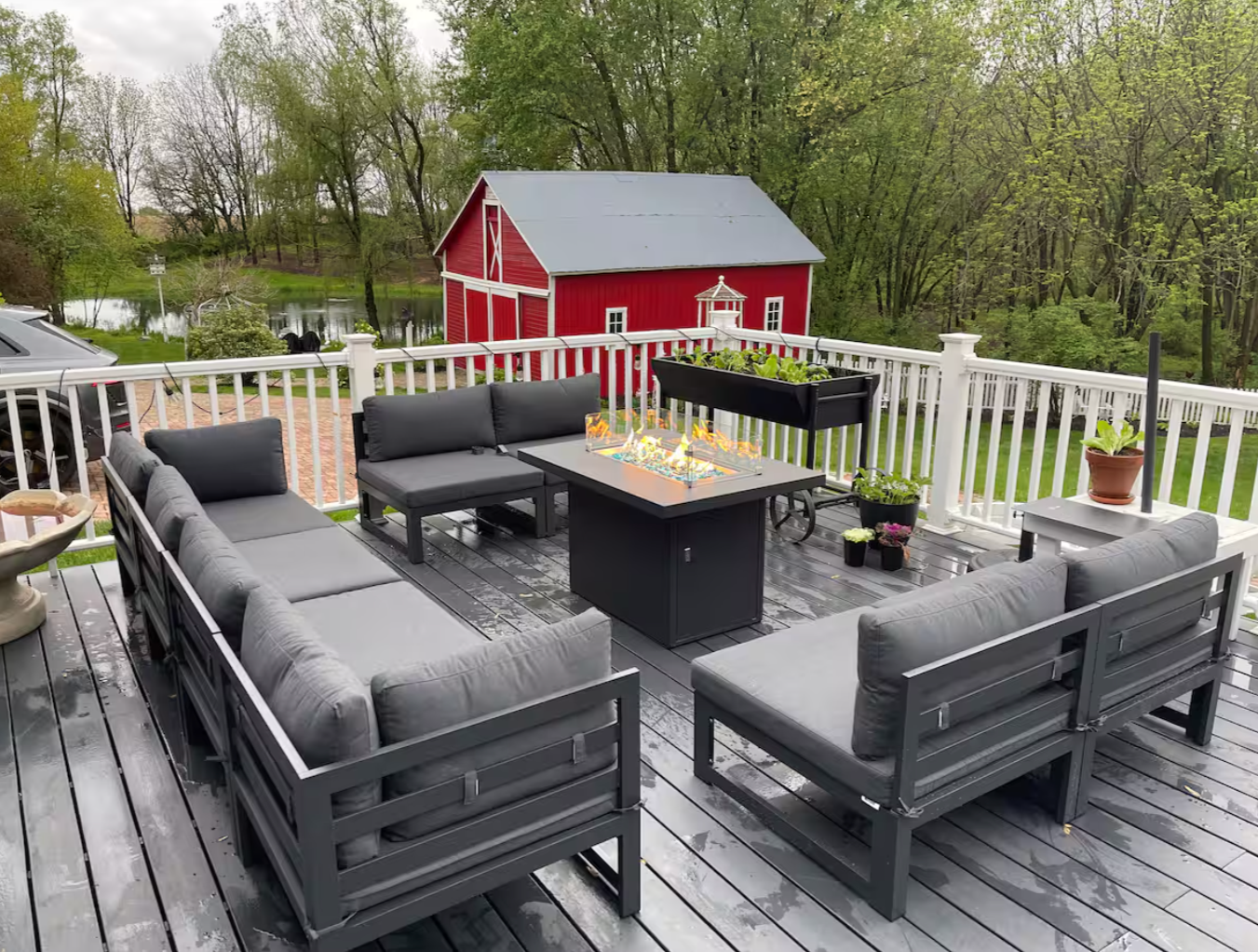 Outdoor deck with a sectional sofa, a fire pit table, potted plants, and a white fence, overlooking a pond and a red barn surrounded by green trees.