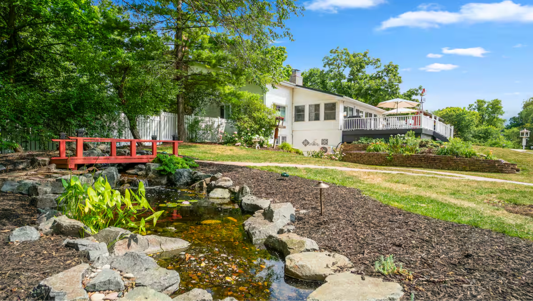 A backyard garden with a small pond surrounded by rocks and green plants, a red bridge over the pond, and a white house with a deck featuring an umbrella in the background under a blue sky with a few white clouds.