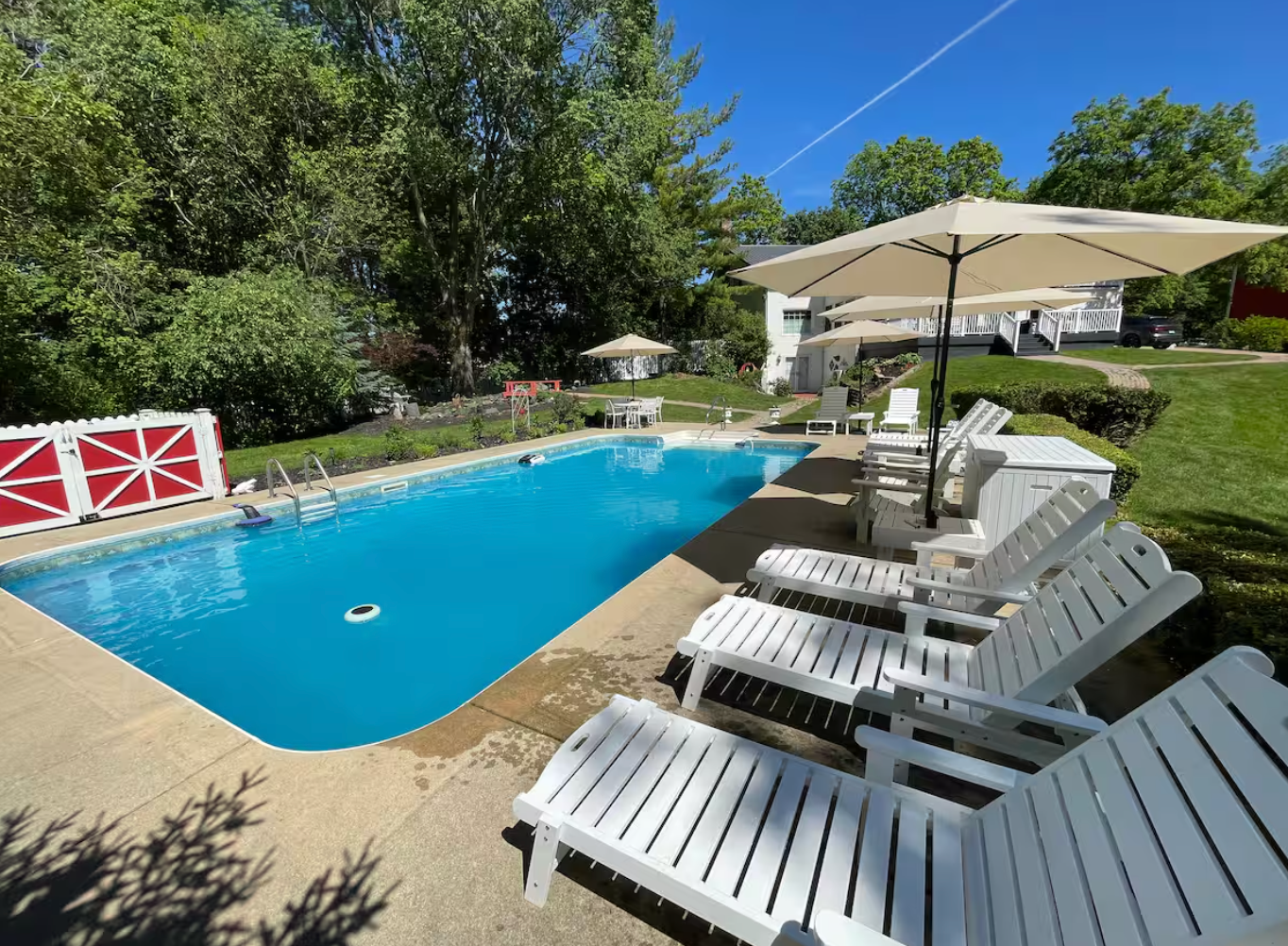 Residential backyard with a rectangular swimming pool, white lounge chairs with umbrellas, surrounded by green lawn and trees, under a clear blue sky.