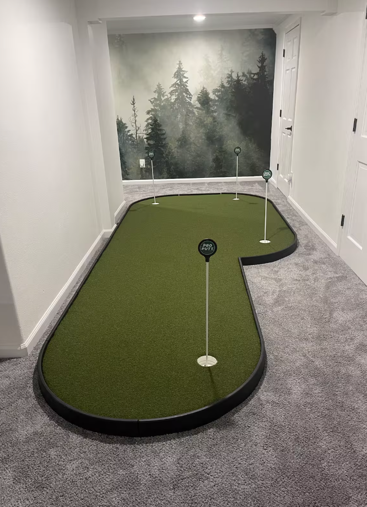 Miniature indoor golf putting green with four flags, one labeled 'Pro Putt,' set up in a room with gray carpet and a forest mural on the back wall.