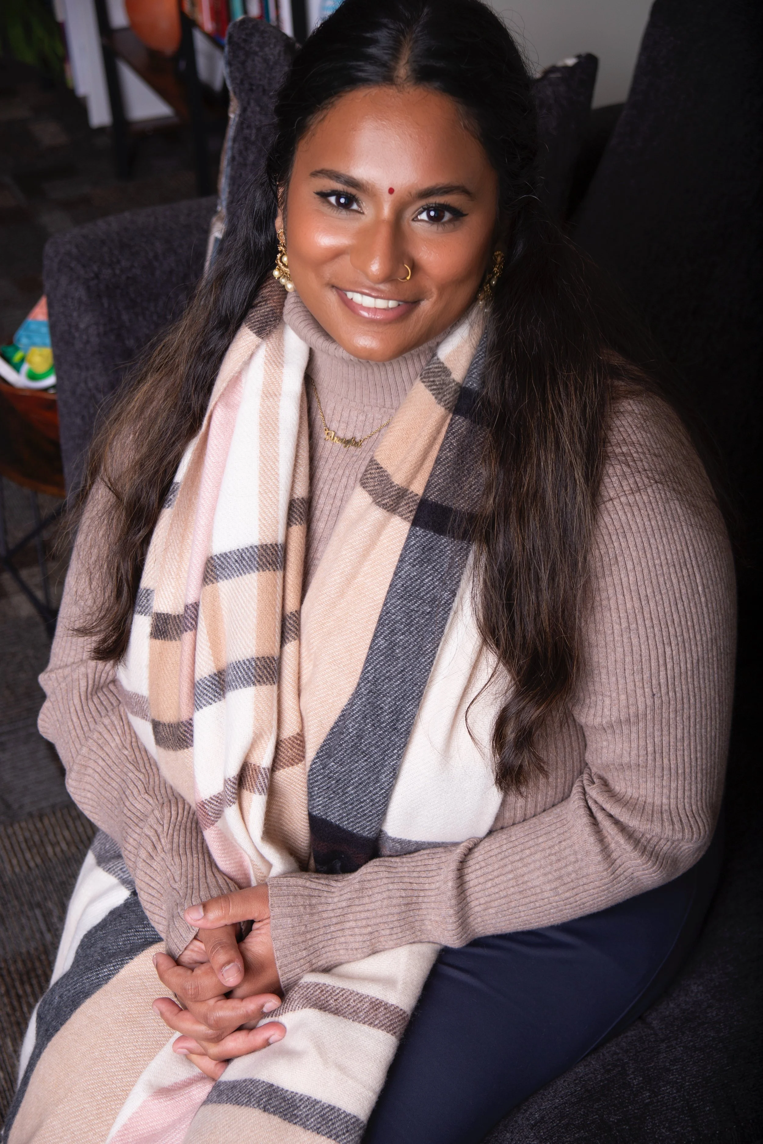 A woman with long dark hair, wearing a beige turtleneck sweater, a plaid scarf, and dark pants, sitting on a black couch and smiling at the camera.