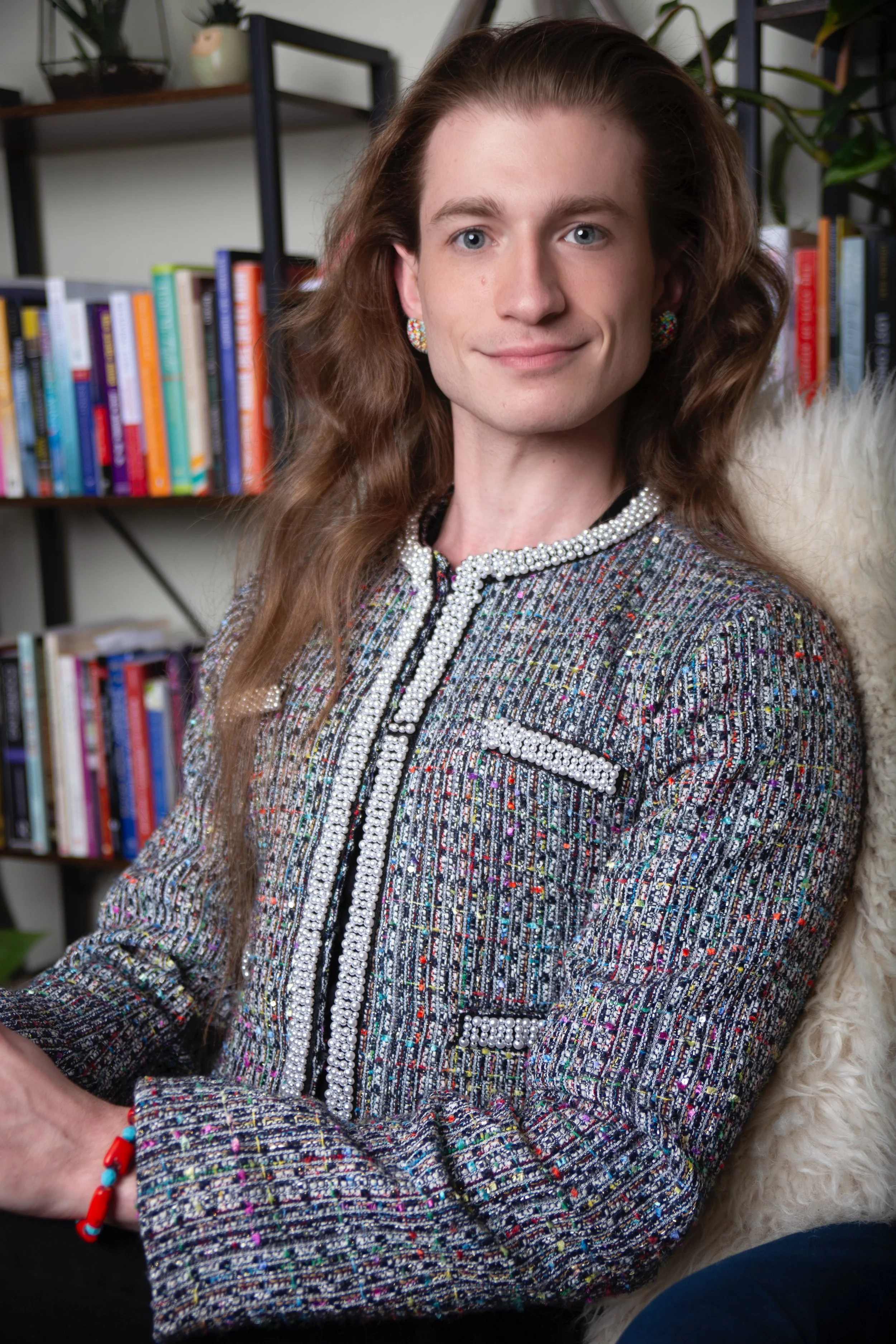 A young woman with long, curly brown hair, sitting indoors in front of a bookshelf filled with colorful books. She is wearing an elegant tweed jacket decorated with pearls, earrings, and a colorful bracelet, and smiling at the camera.
