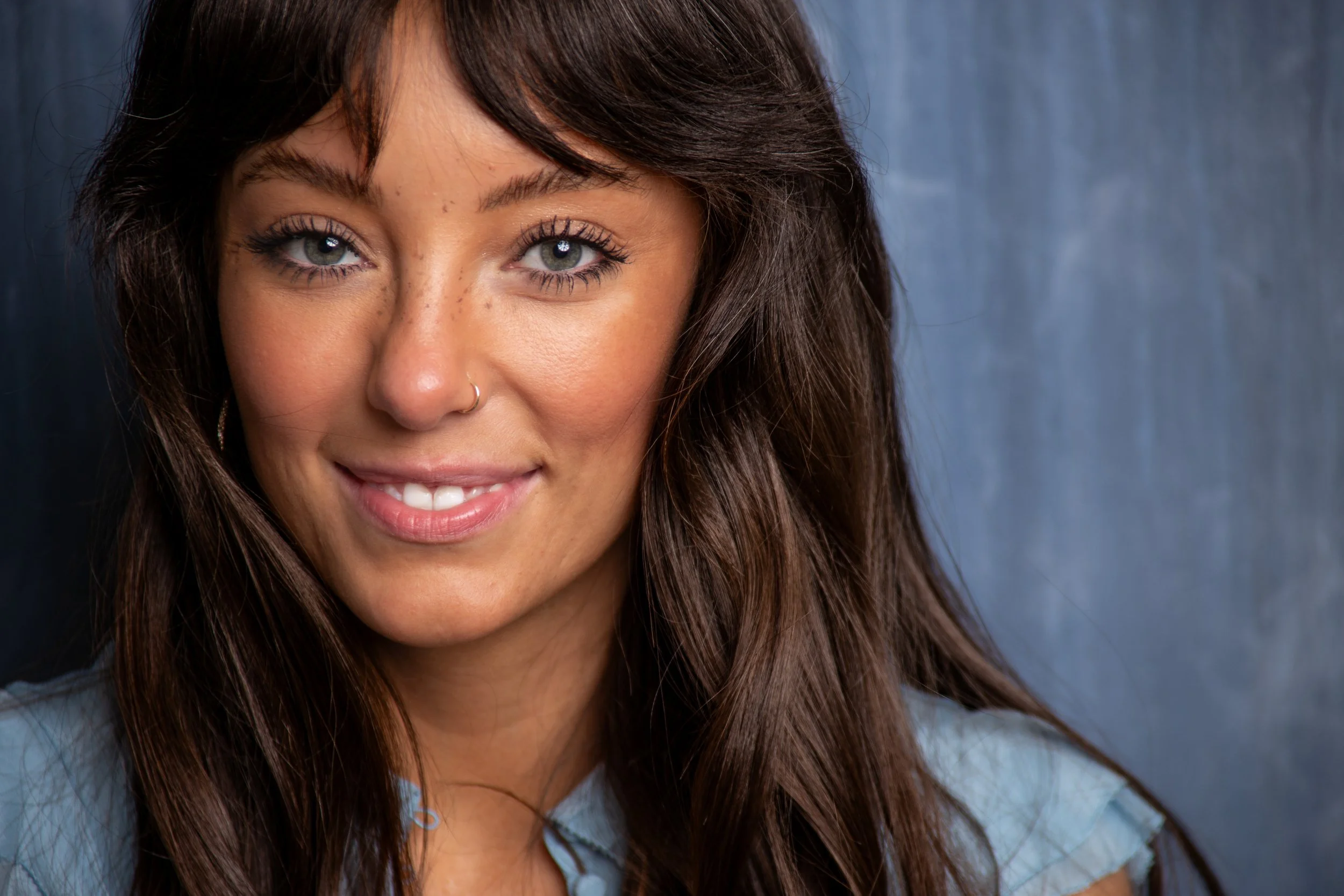 A young woman with long brown hair, freckles, gray eyes, a nose piercing, and wearing a light blue top, smiling at the camera against a blue background.