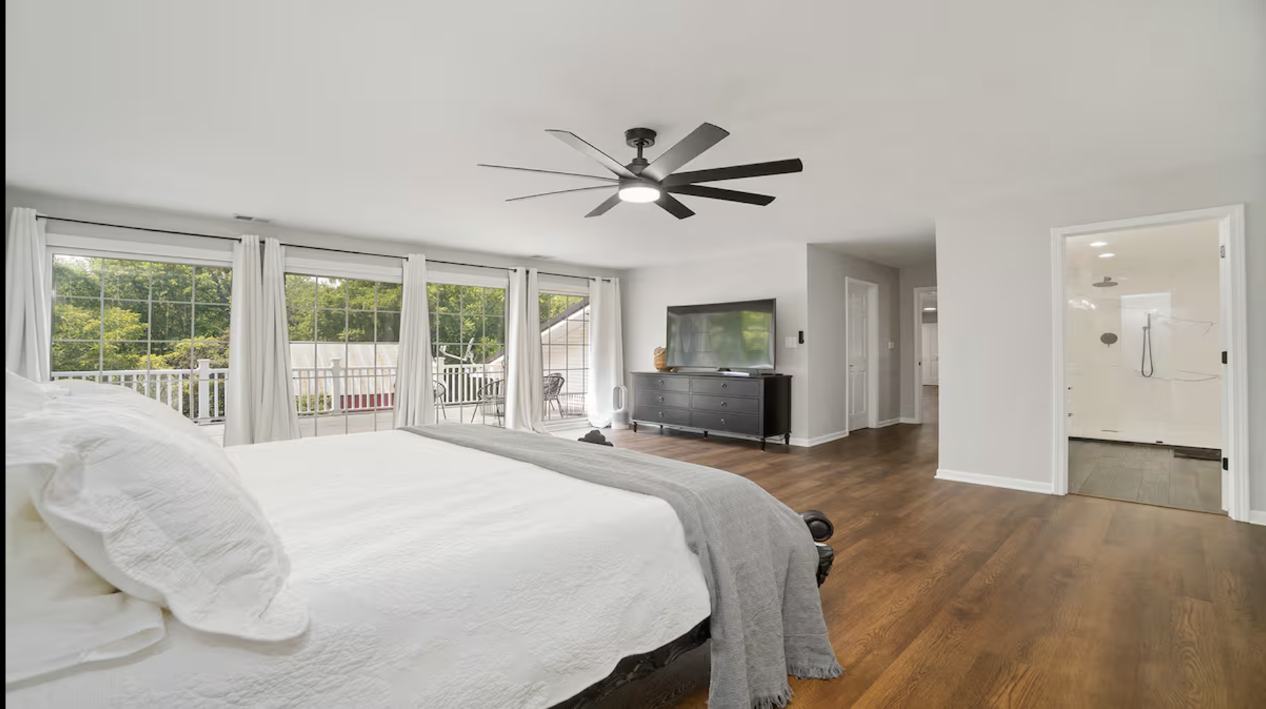 A bedroom with a large bed covered in white bedding and pillows. Large sliding glass doors with white curtains lead to a balcony with outdoor seating. There is a ceiling fan with multiple blades. A black dresser with a flat-screen TV on top is against the wall. The floor is wooden, and an open doorway reveals a bathroom with a walk-in shower.
