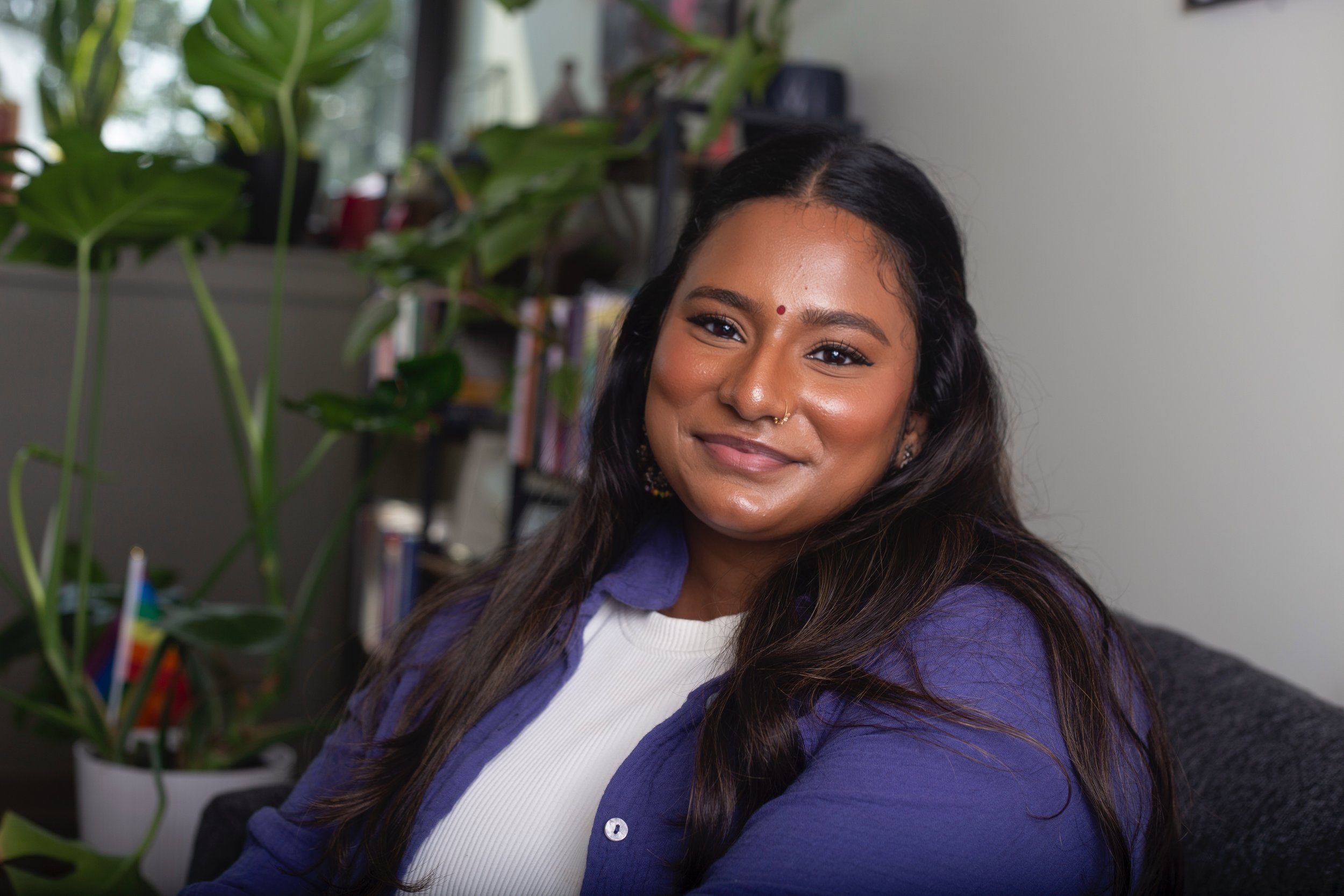 A smiling woman with long dark hair wearing a white shirt and purple jacket, sitting indoors with houseplants and books in the background.