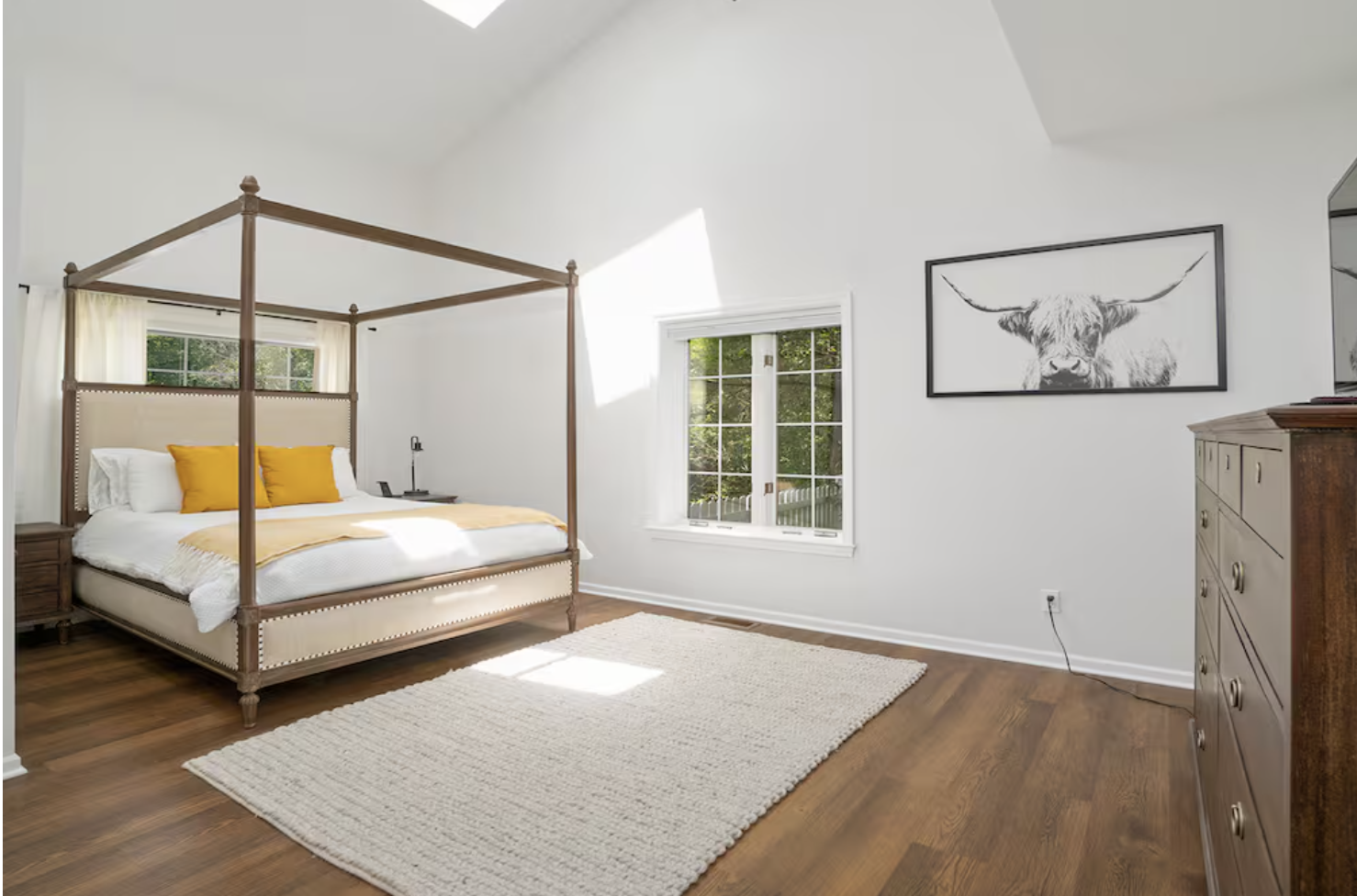 Bright bedroom with a four-poster bed, white bedding, yellow pillows, wooden nightstand, and dresser, with a large window and a framed black-and-white cow portrait on the wall.