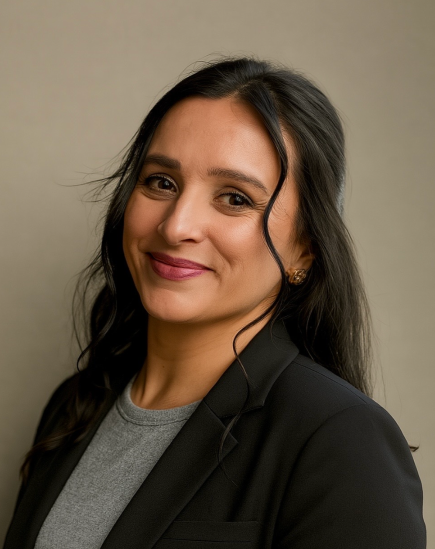 Professional woman with dark wavy hair and earrings, smiling in a black blazer and gray top against a neutral background.