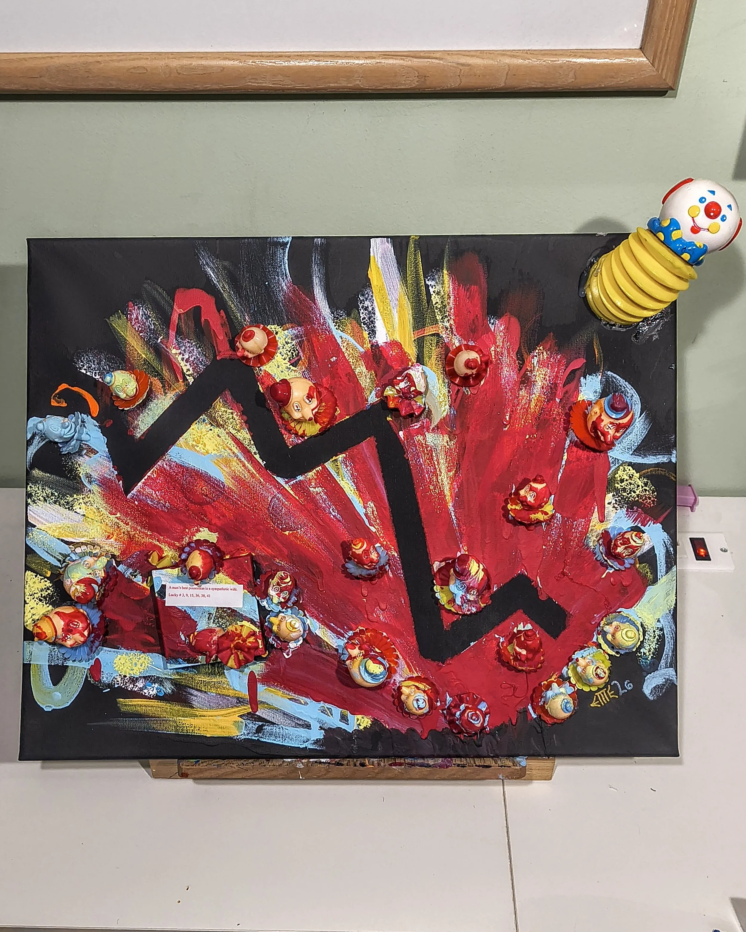 A colorful abstract painting with a black zigzag line, red, yellow, white, and blue splashes, and small clown figurines attached across the artwork.