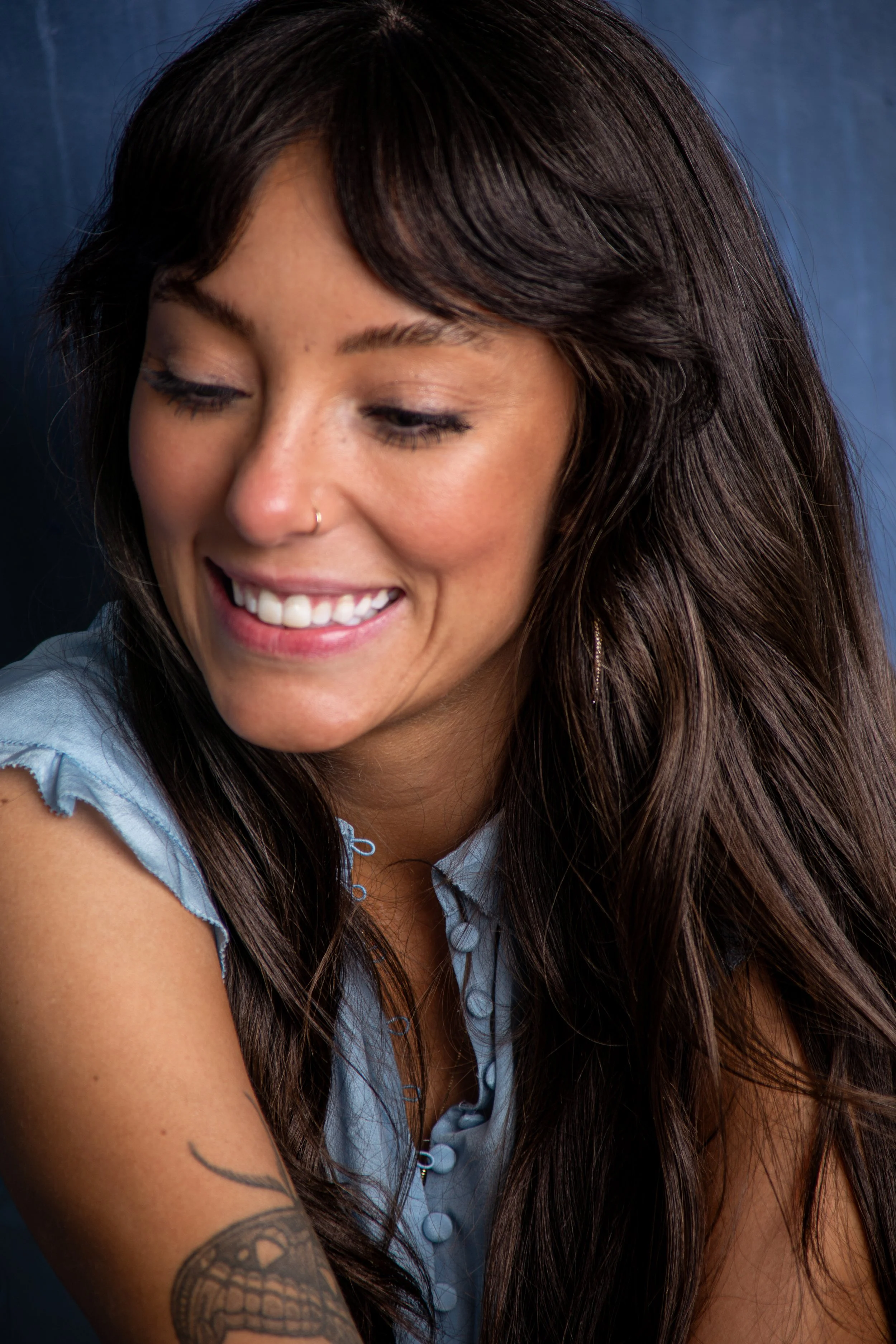 Close-up photo of a smiling woman with long brown hair, a nose piercing, wearing a light blue top, and showing a tattoo on her arm, against a dark background.