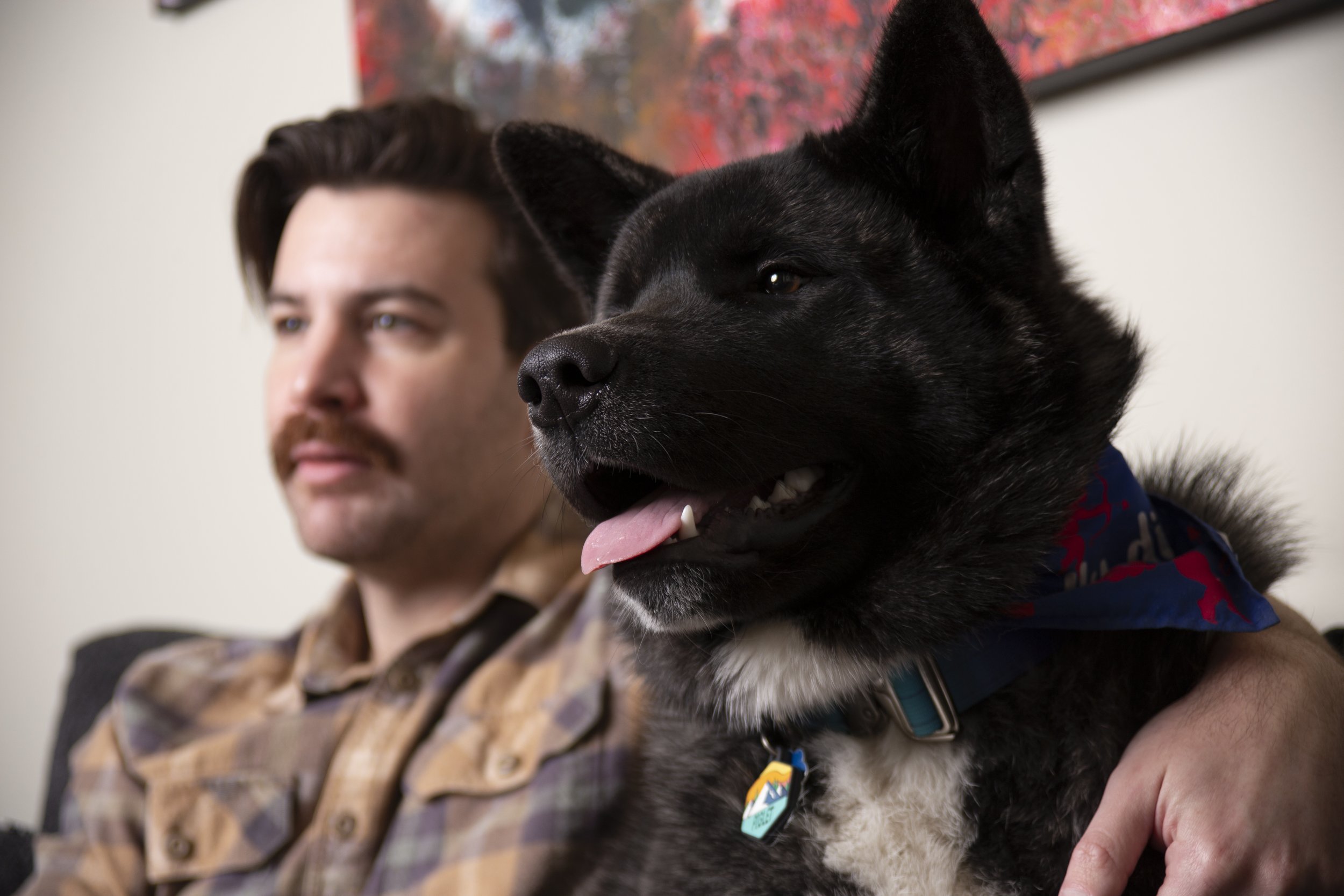 A man with brown hair and a mustache sitting on a couch with a black and white dog. The man is looking to his left, and the dog, wearing a blue bandana, is sitting close to him with its tongue out.