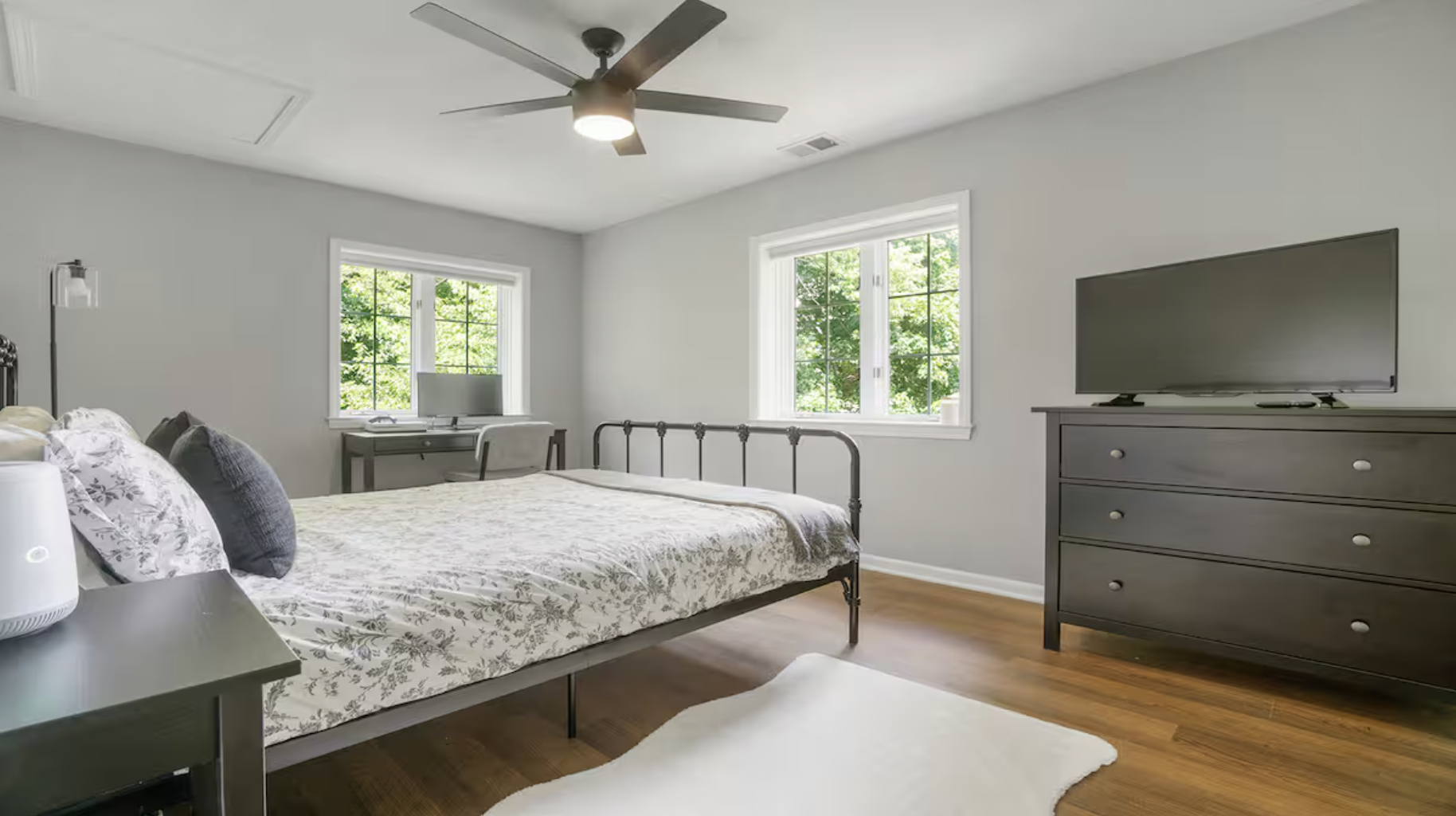 A bedroom with a metal bed frame, floral bedding, and three pillows, a white rug on wooden floor, a dresser with a flat-screen TV, and a small desk with a computer and chair near two windows showing green foliage, a ceiling fan with four blades, and white walls.