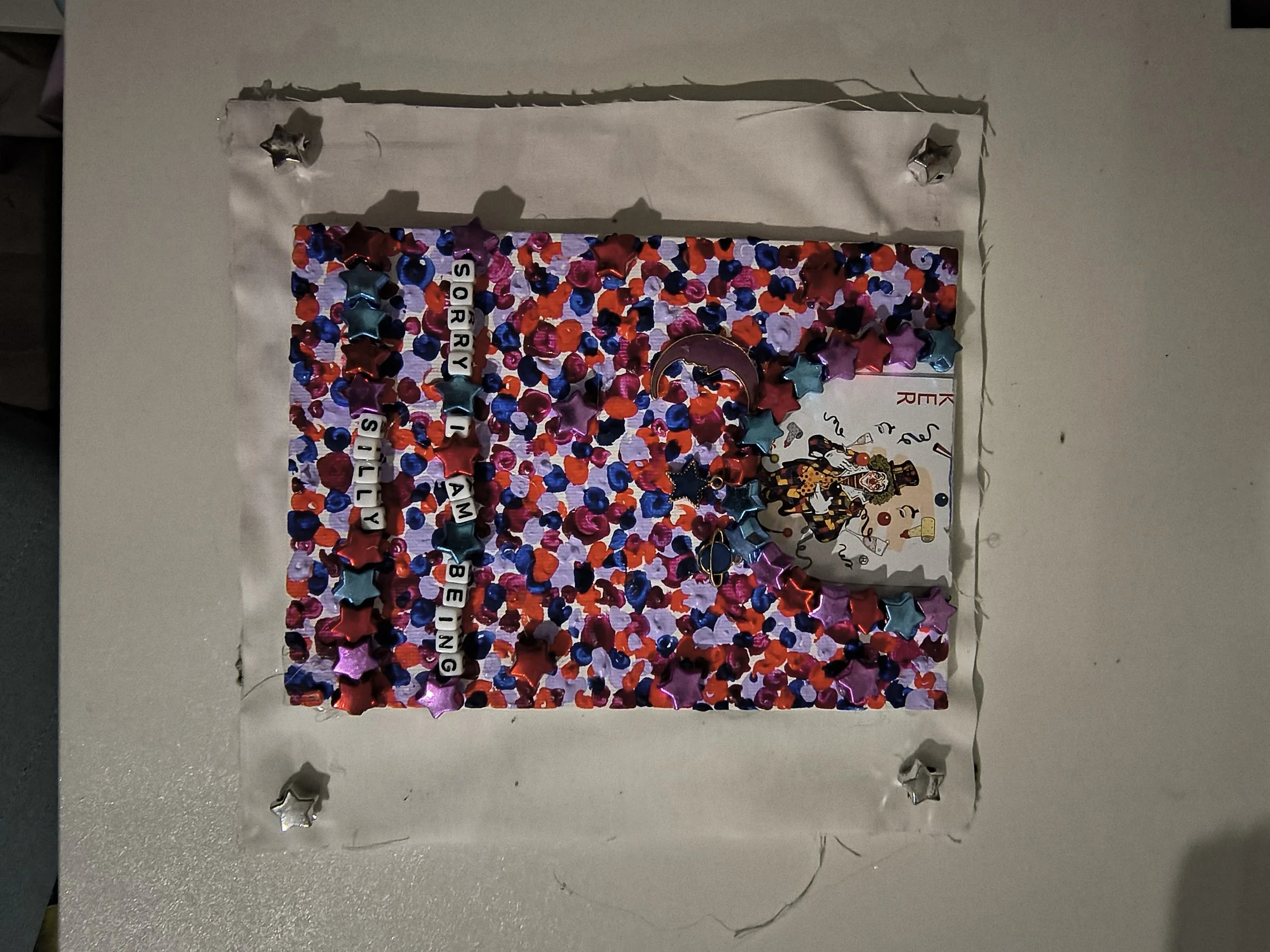 A decorated bulletin board with multicolored confetti, star-shaped metallic decorations, and a message spelling out "Sorry I am being silly" made with letter beads. There is a partial picture of a clown or jester and some paper decorations on the rig