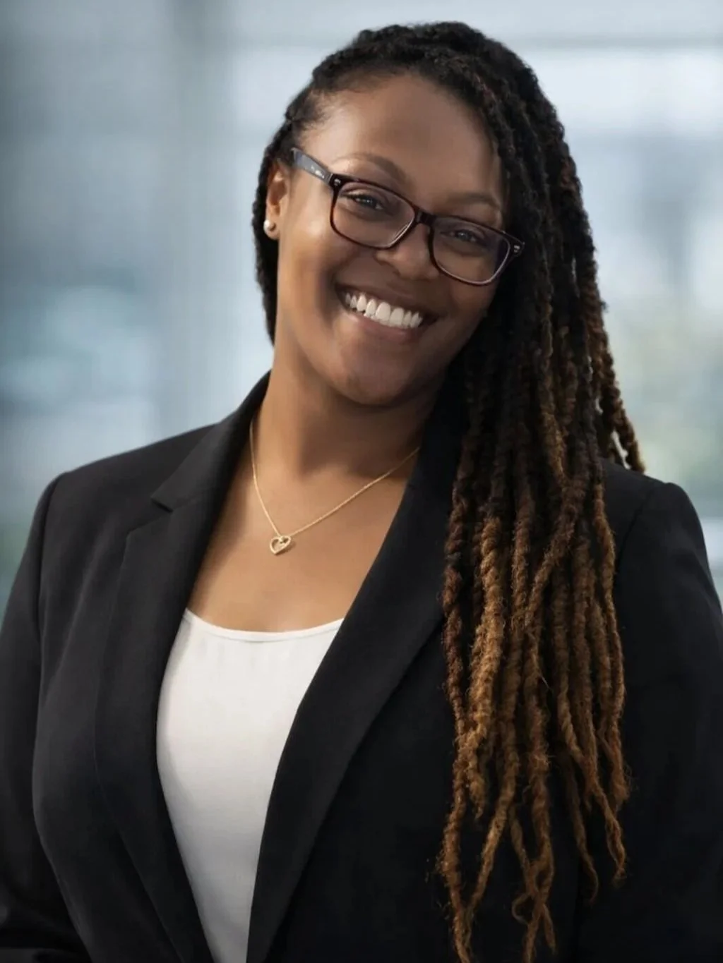 A smiling woman with glasses and long dreadlocks, wearing a black blazer and white top, standing in an indoor setting.