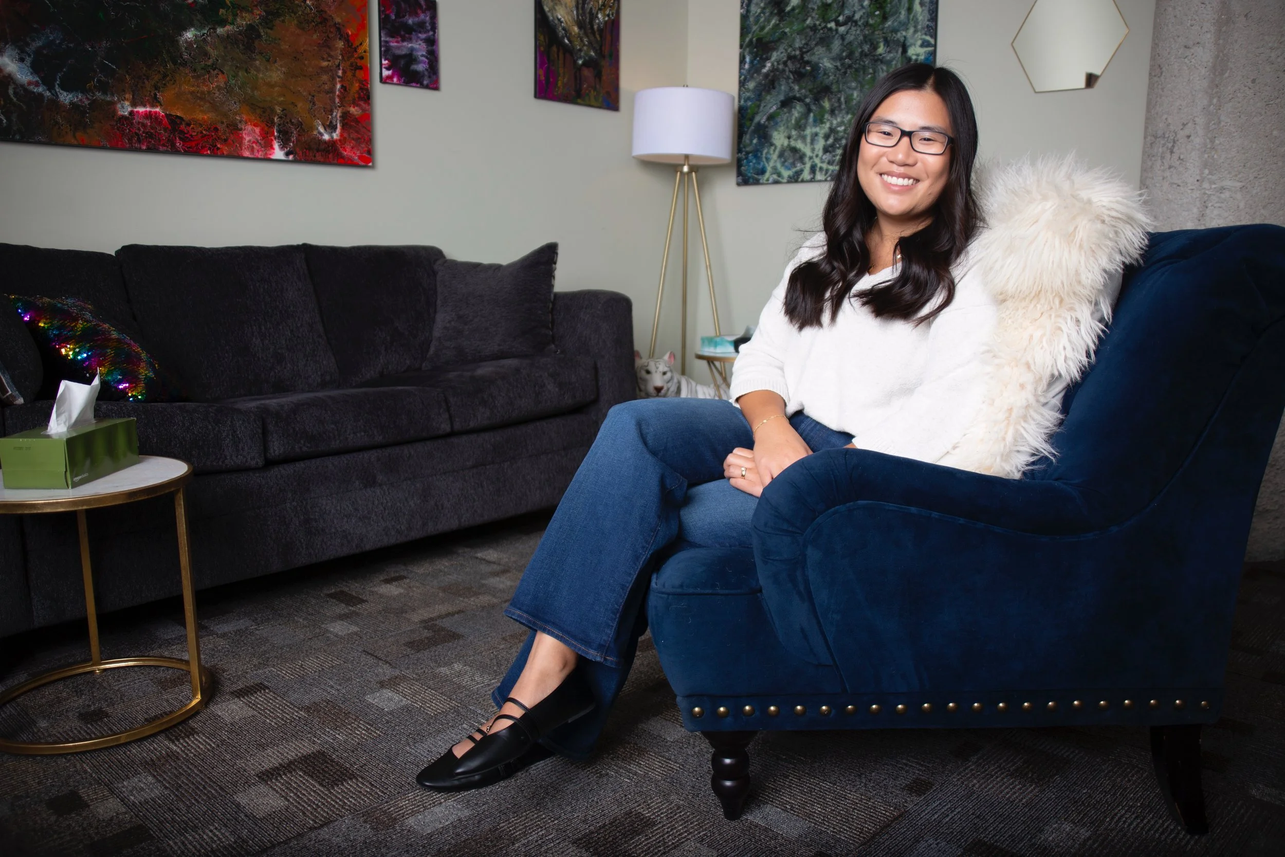 A woman with glasses, long dark hair, wearing a white sweater and blue jeans, sitting on a dark blue armchair with a fluffy white pillow, smiling at the camera in a modern living room with colorful artwork on the wall, a black sofa, a small round side table with tissues, and a white and gray cat in the background.