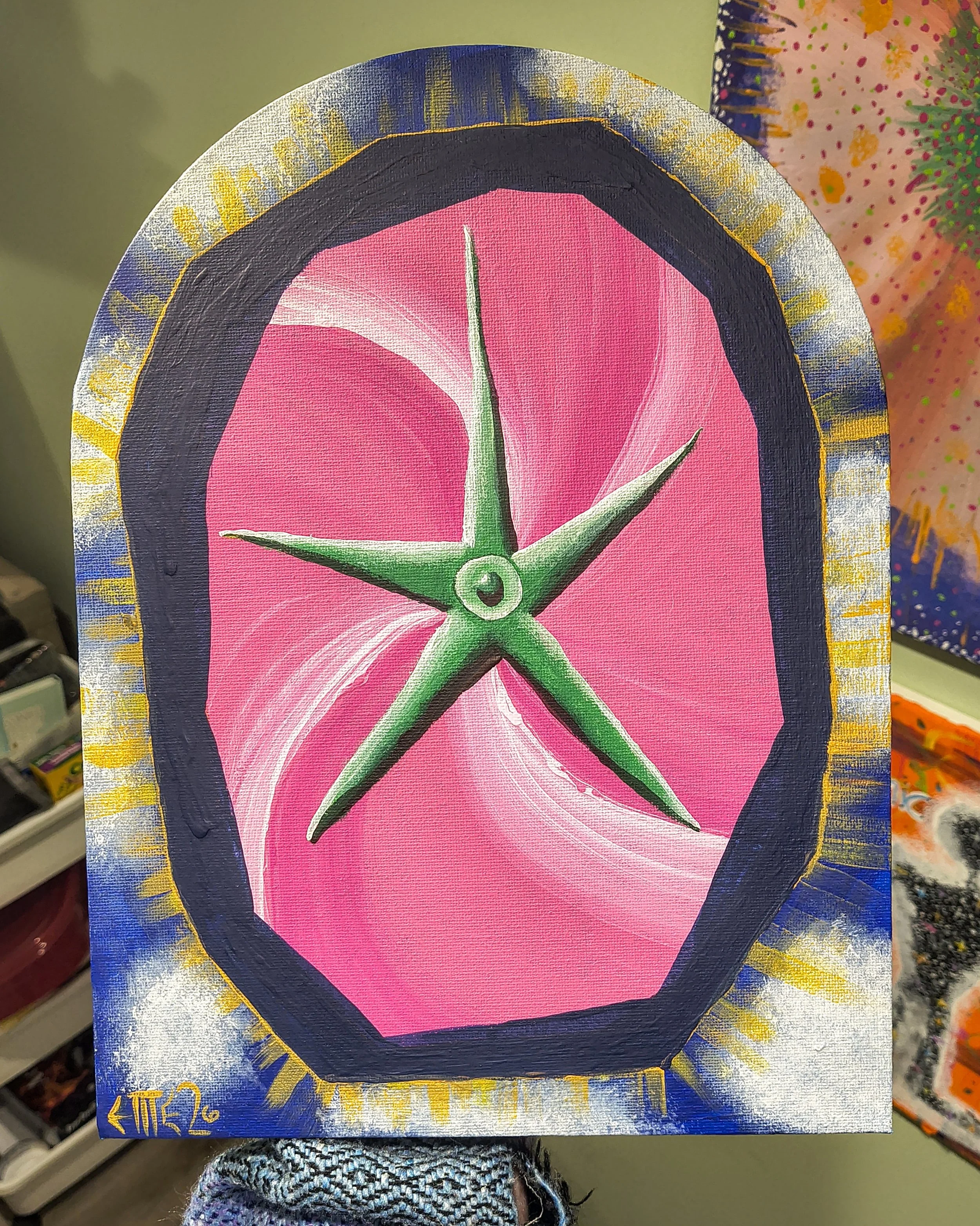 A painting of a green star with five points set against a pink background inside a black-bordered oval. The painting has a colorful decorative border with blue, yellow, and white accents.