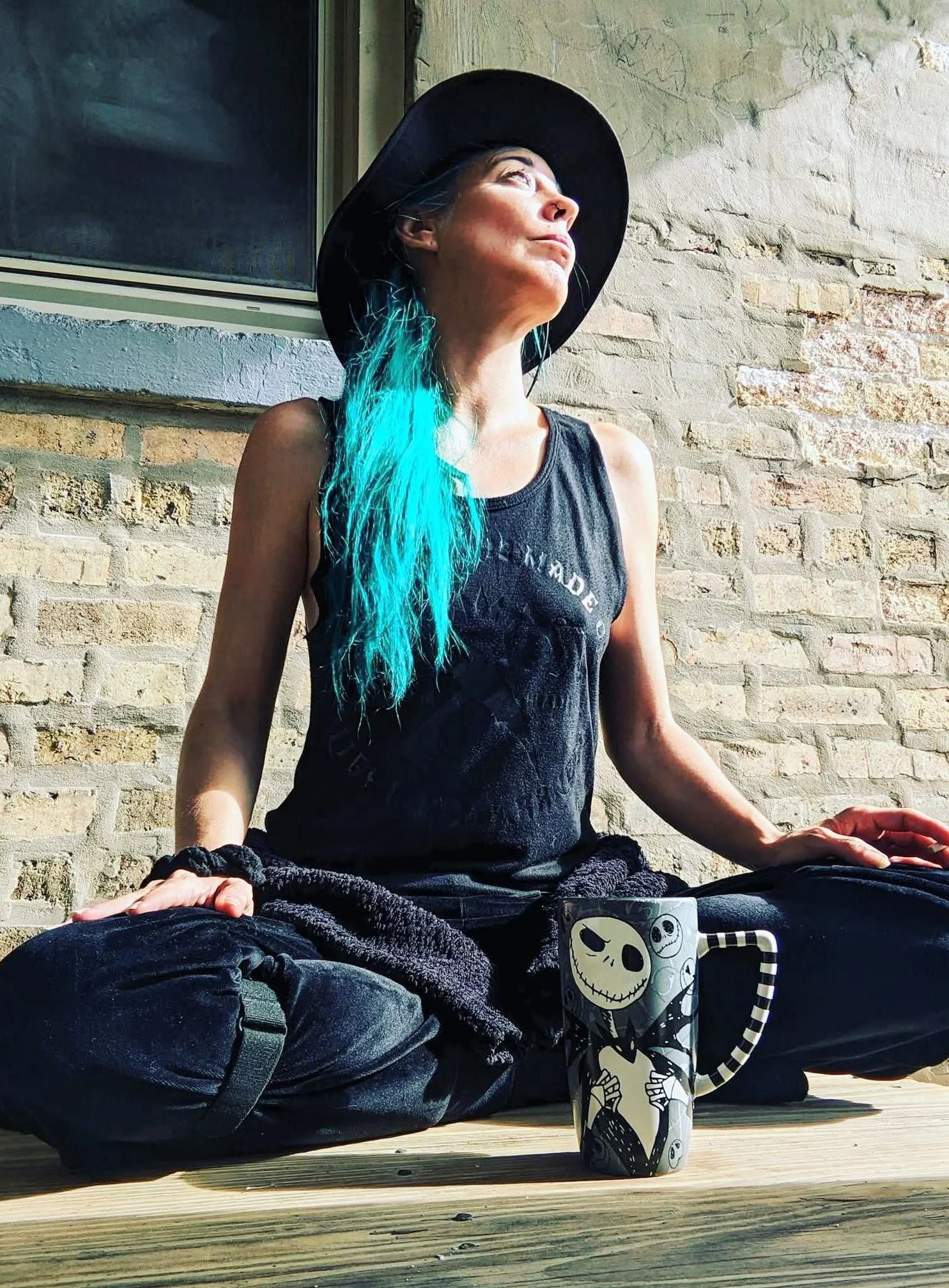 A woman with teal-dyed hair, wearing a black wide-brimmed hat and a sleeveless black shirt, is sitting cross-legged on a wooden deck, appearing to meditate. A ceramic mug decorated with a skeleton design and a striped straw is in front of her. She is outdoors near a brick wall and window, looking upward with a calm expression.