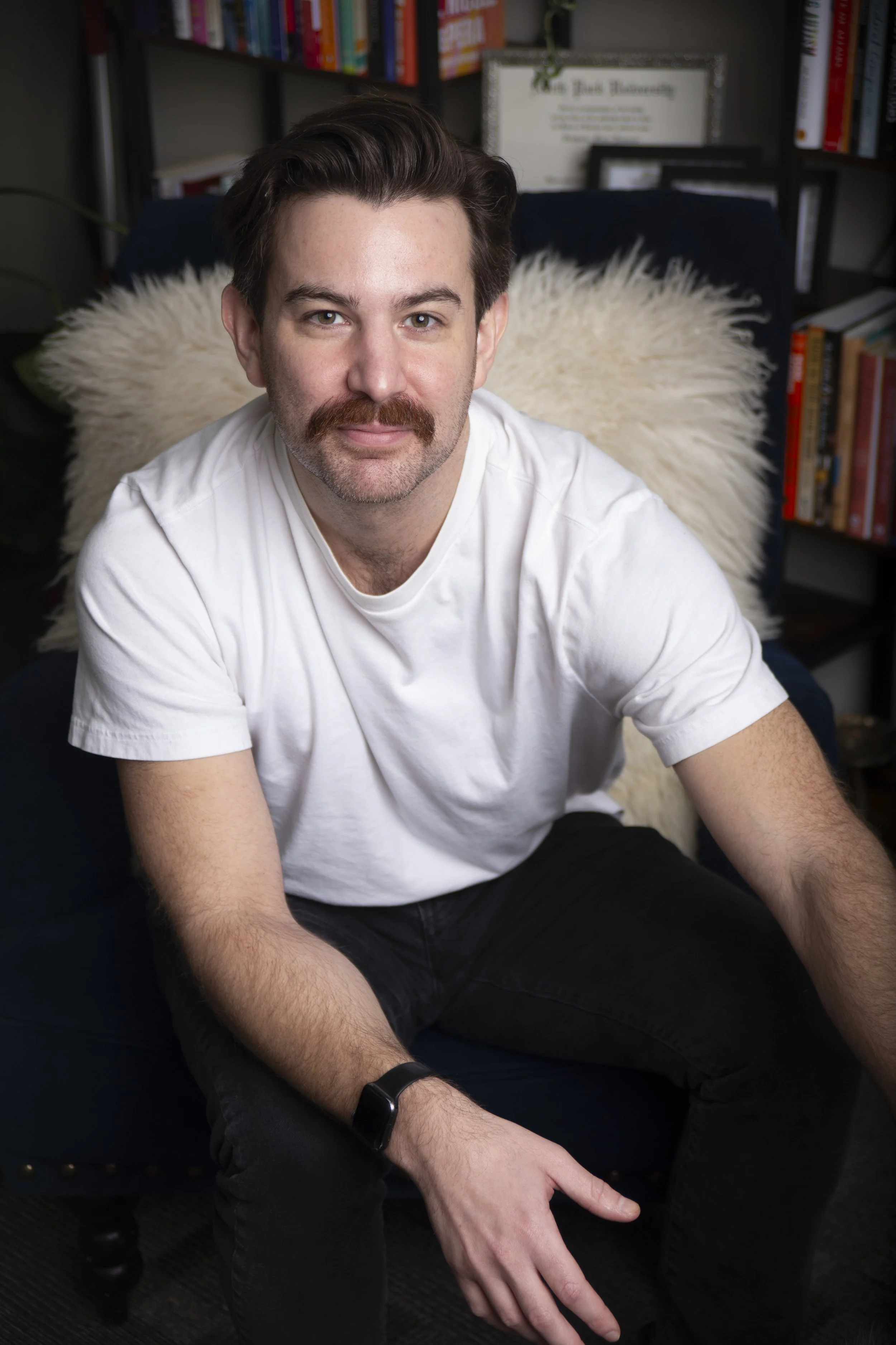 A man with a mustache and beard, wearing a white t-shirt, sits on a dark blue chair in a room with bookshelves in the background.