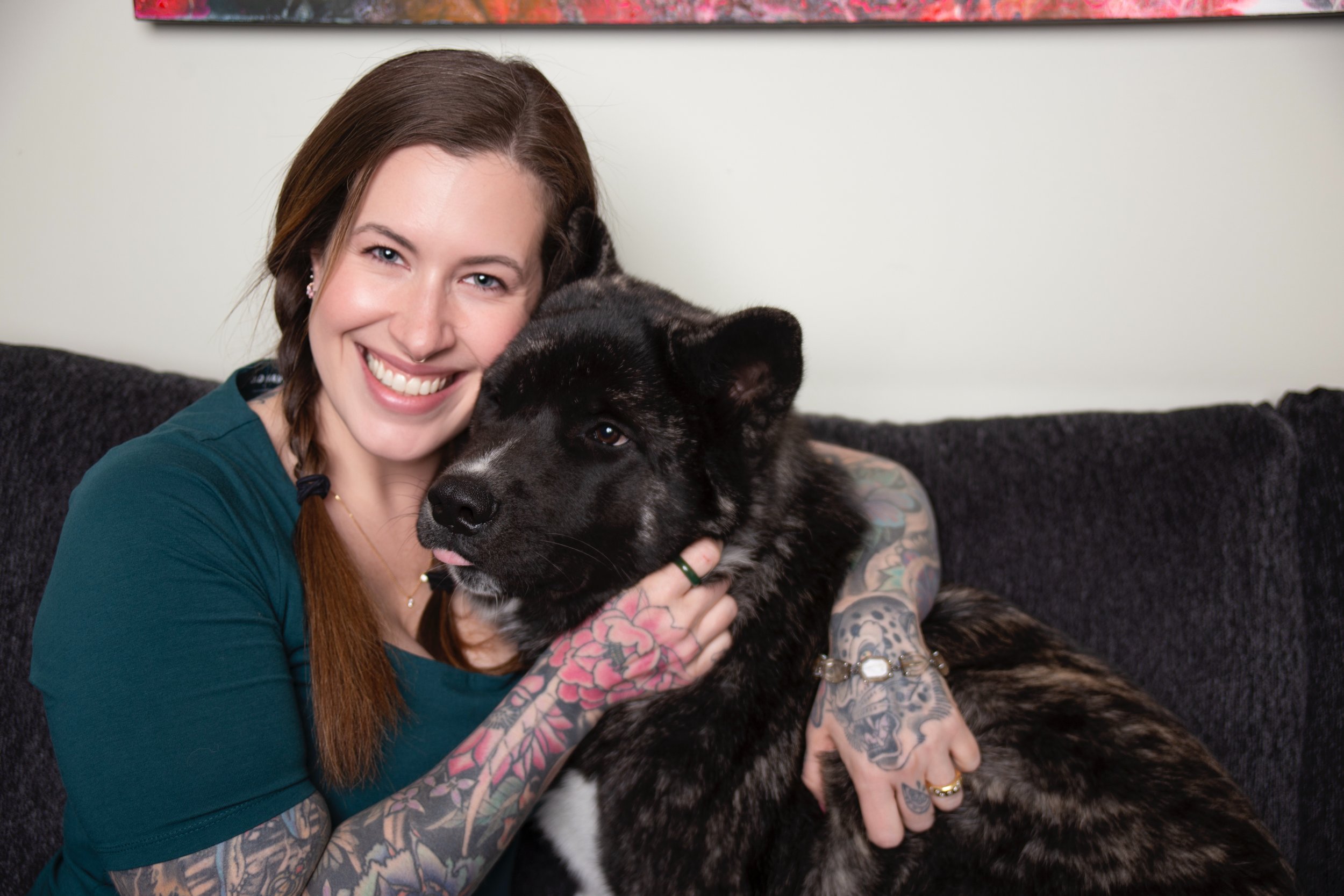 A young woman with long brown hair, tattoos on her arms, wearing a teal shirt and jewelry, smiling while hugging a black and white dog with a fluffy coat and a pink tongue sticking out, sitting on a dark couch.