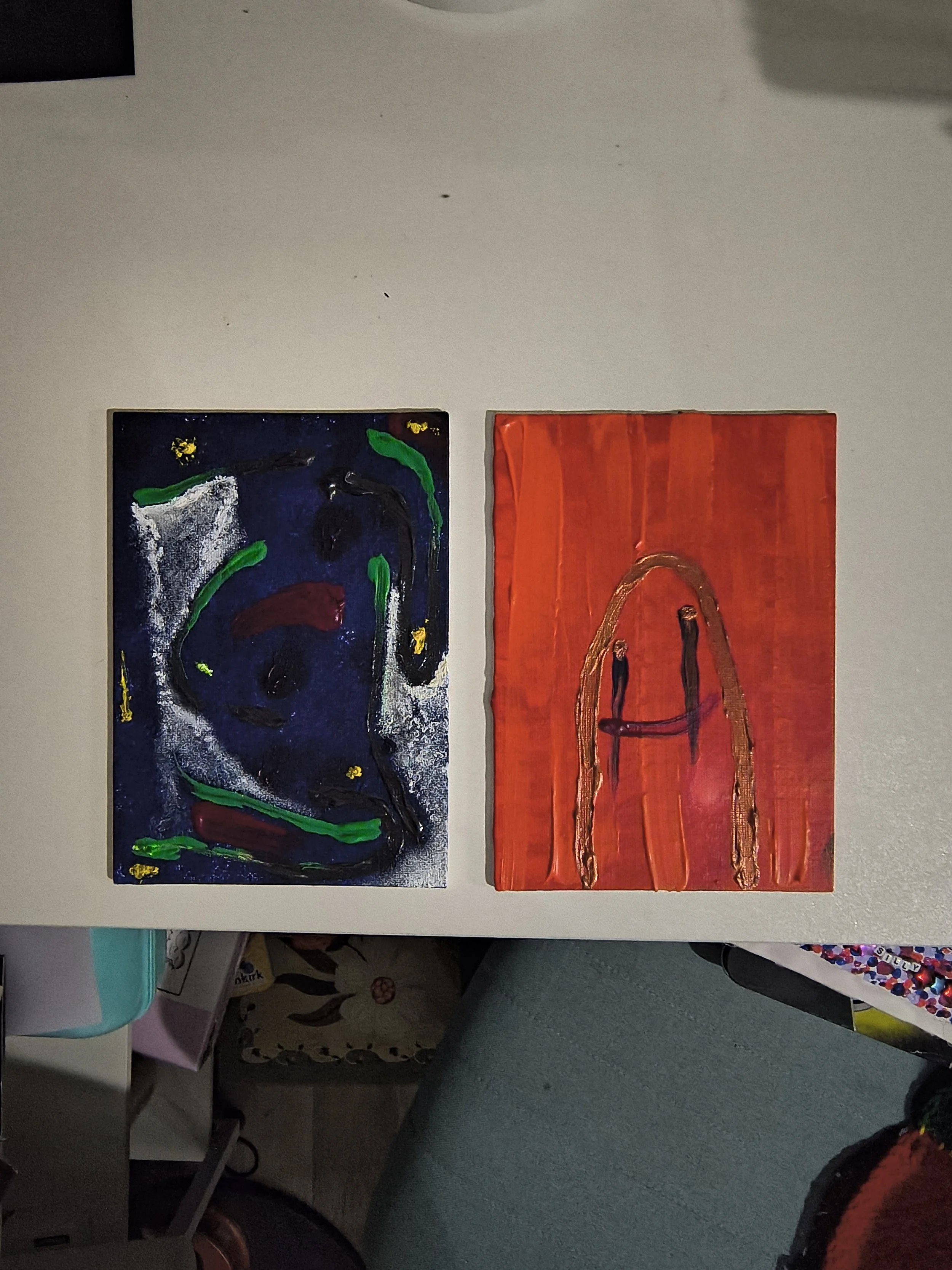 Two abstract paintings side by side on a white table, one with dark blue, green, white, red and yellow colors and the other with red background and brown and black lines.