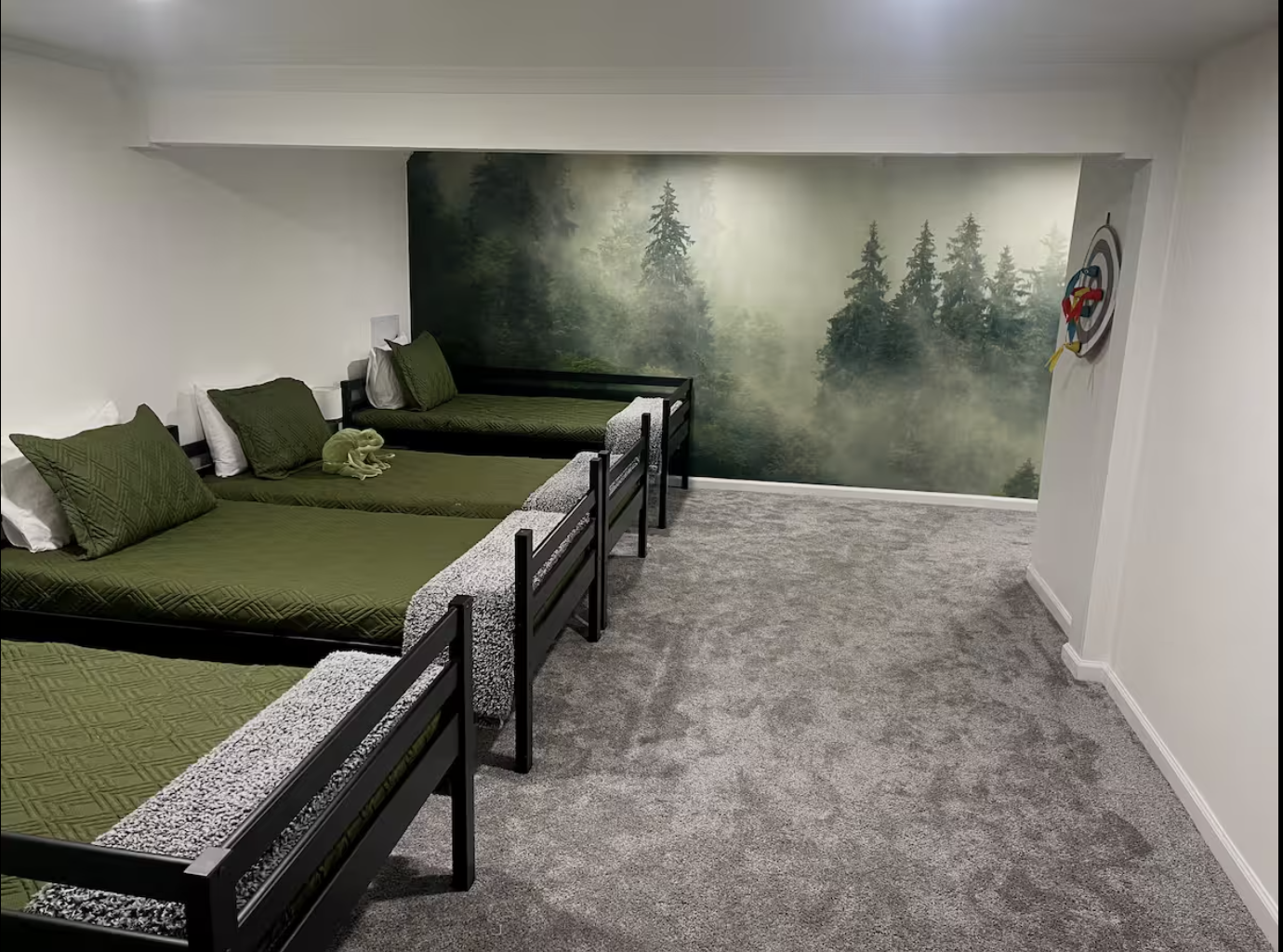 Three twin beds with green bedding and pillows, a forest-themed mural on the back wall, and a small decorative target on the white wall.