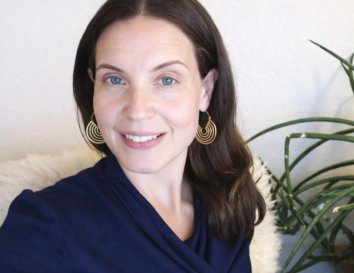 A woman with fair skin and long brown hair, wearing large gold spiral earrings and a dark blue top, smiling while sitting on a white fur-covered chair next to a green plant.