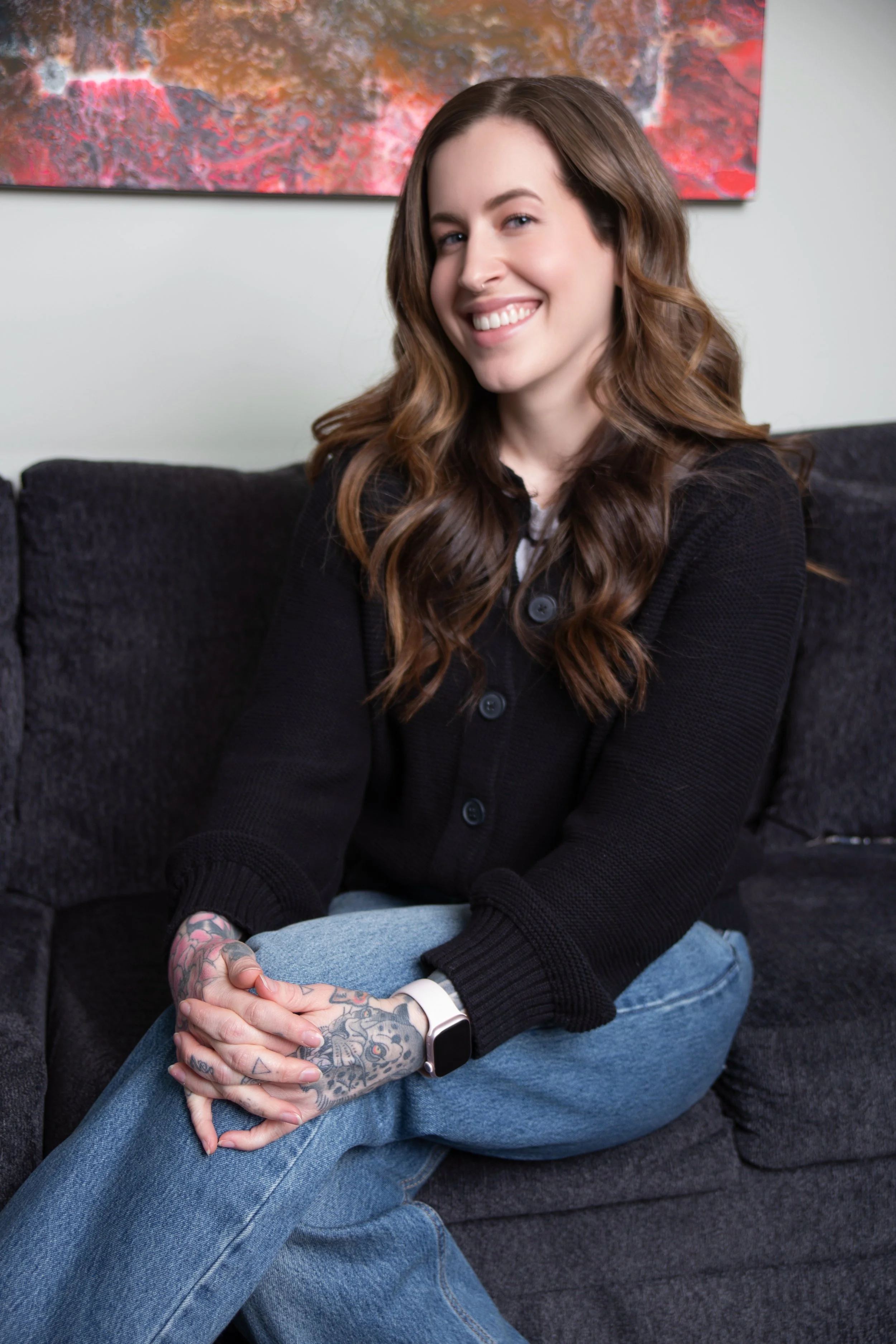 A woman with long, wavy brown hair, smiling, sitting on a black couch with her legs crossed. She is wearing a black cardigan, blue jeans, a smartwatch, and has tattooed hands. There is a colorful abstract painting on the wall behind her. Sex therapy