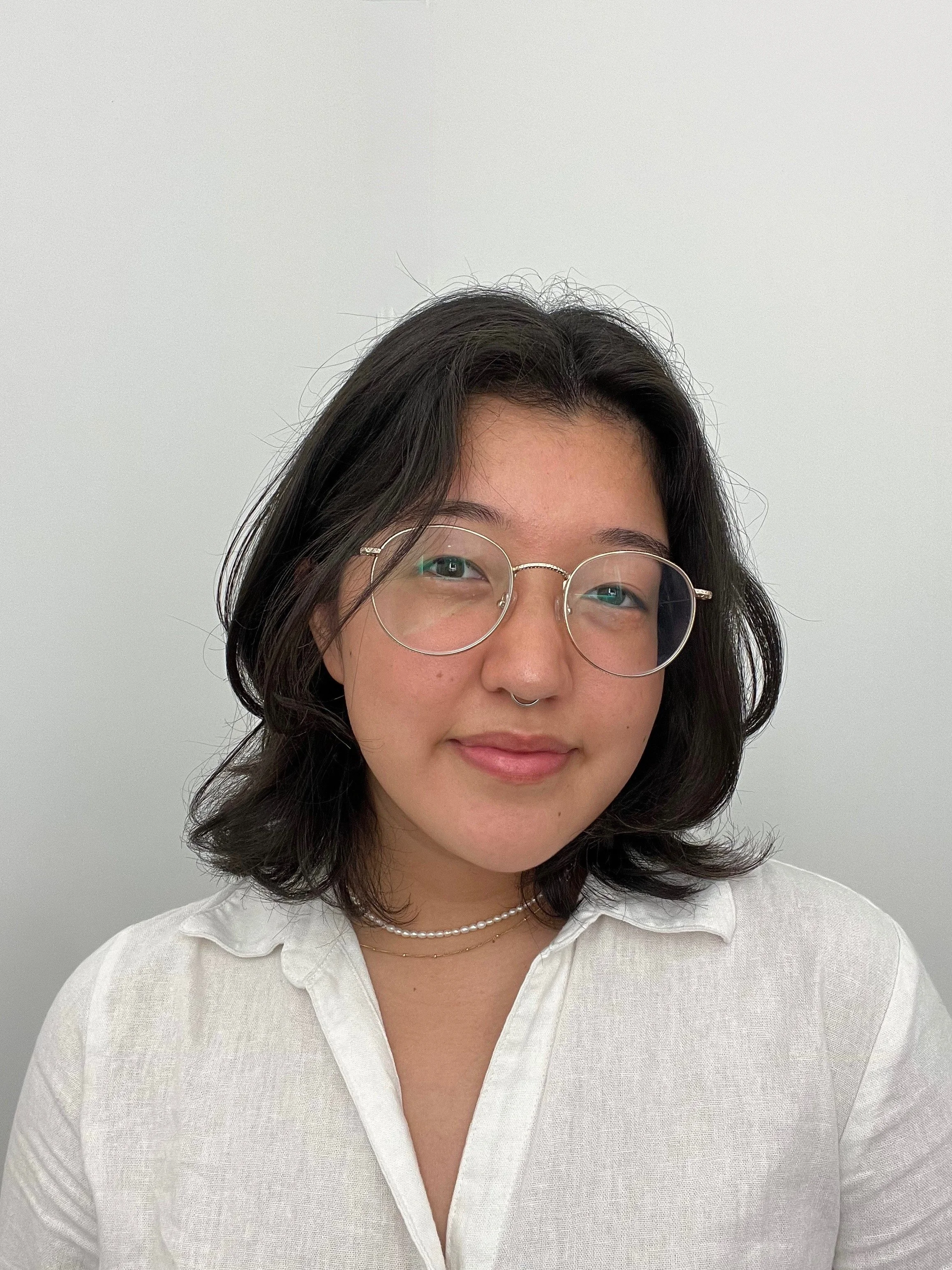 A young woman with shoulder-length black hair, wearing round glasses, a septum piercing, layered necklaces, and a white collared shirt, smiling softly against a plain white background.