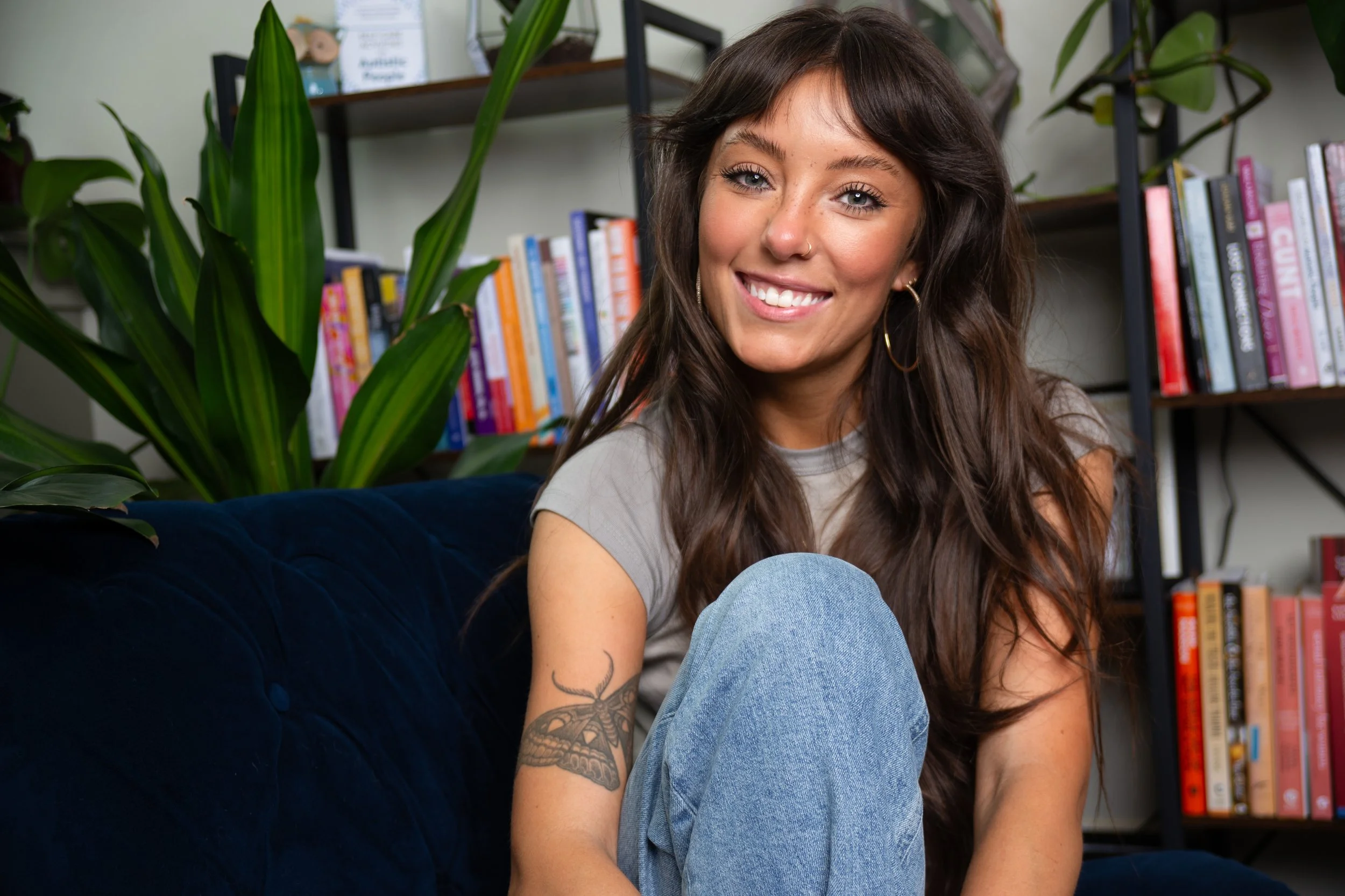 A young woman with long brown hair, freckles, and a nose ring smiling indoors, near a bookshelf and green plants.