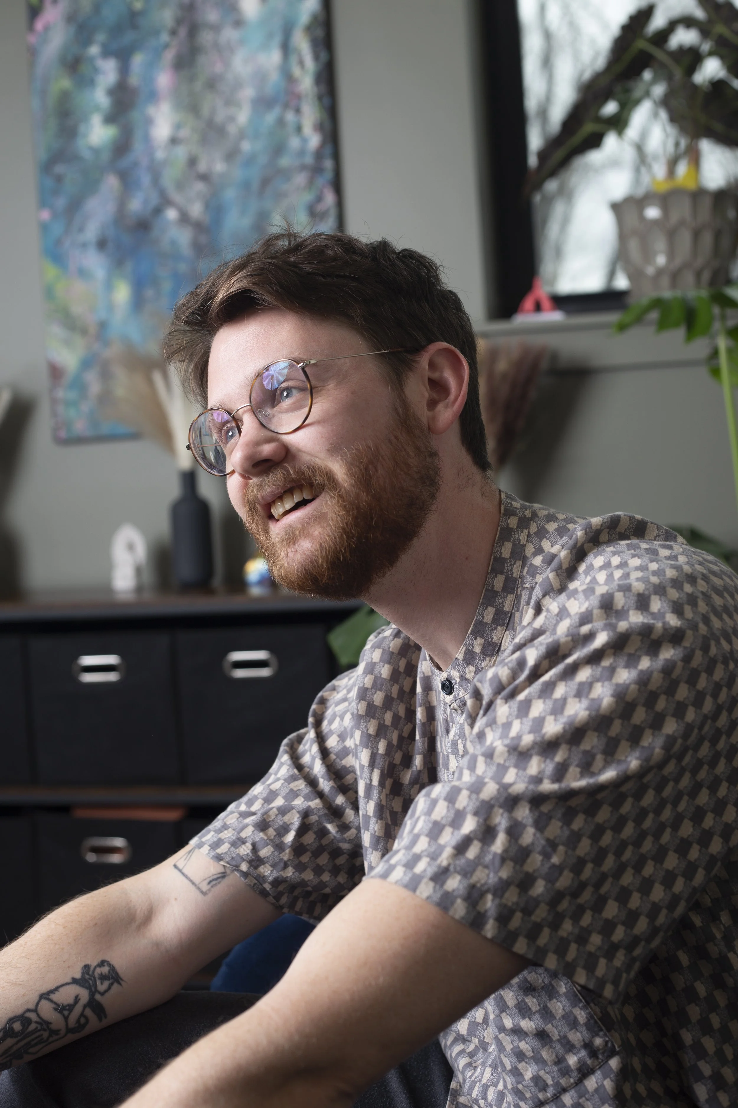 A young man with glasses and a beard is smiling while sitting indoors. He is wearing a checkered shirt, and has tattoos on his arms. Background includes a colorful abstract painting, a window with a view of plants outside, and indoor plants.