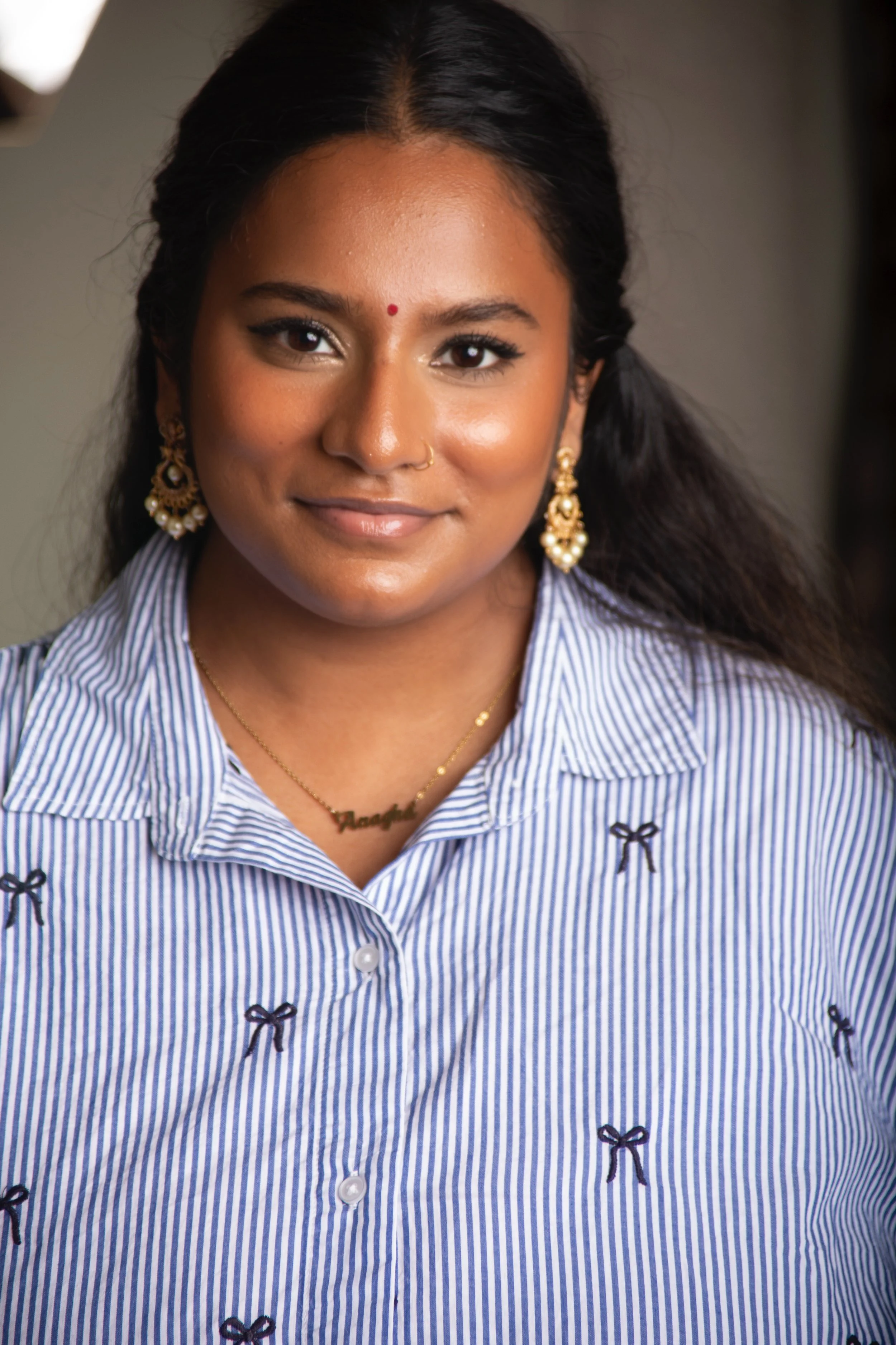 A young woman with long black hair, wearing gold earrings, a necklace, and a blue and white striped blouse with small black bow accents, smiling at the camera.