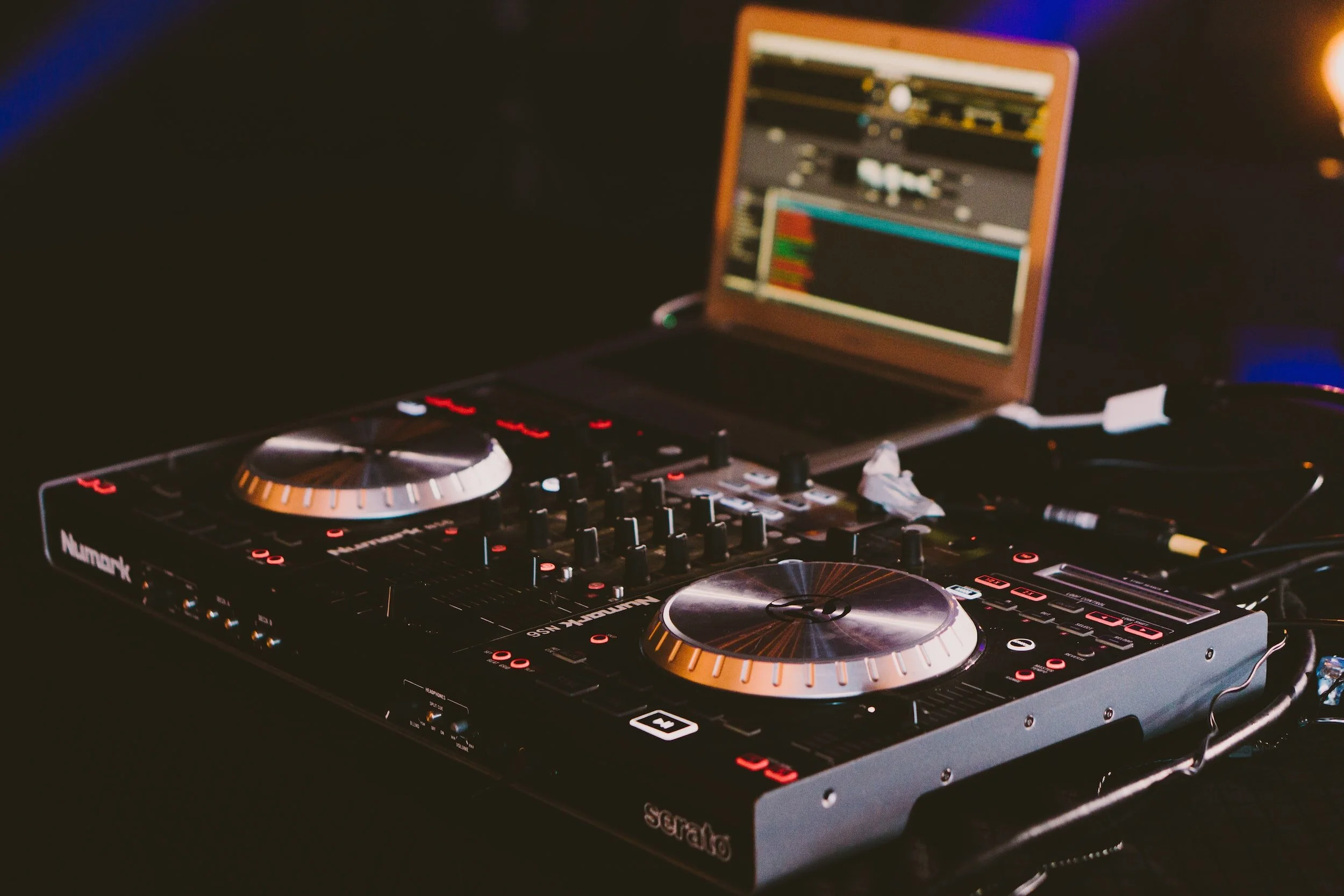 DJ mixing console with a laptop in the background, set up in a dark environment with colorful lighting.