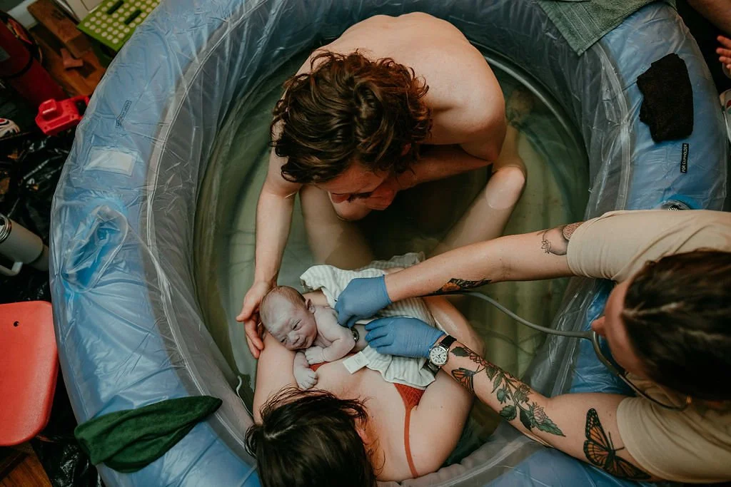 A newborn baby being gently bathed in an inflatable birthing pool by medical personnel and the mother, with a support person also present.