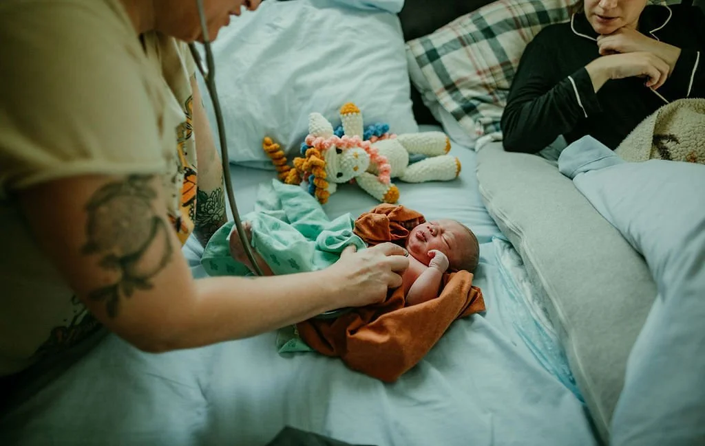 A healthcare worker with tattoos checks on a newborn baby on a hospital bed, with a woman observing, and a stuffed rabbit nearby.